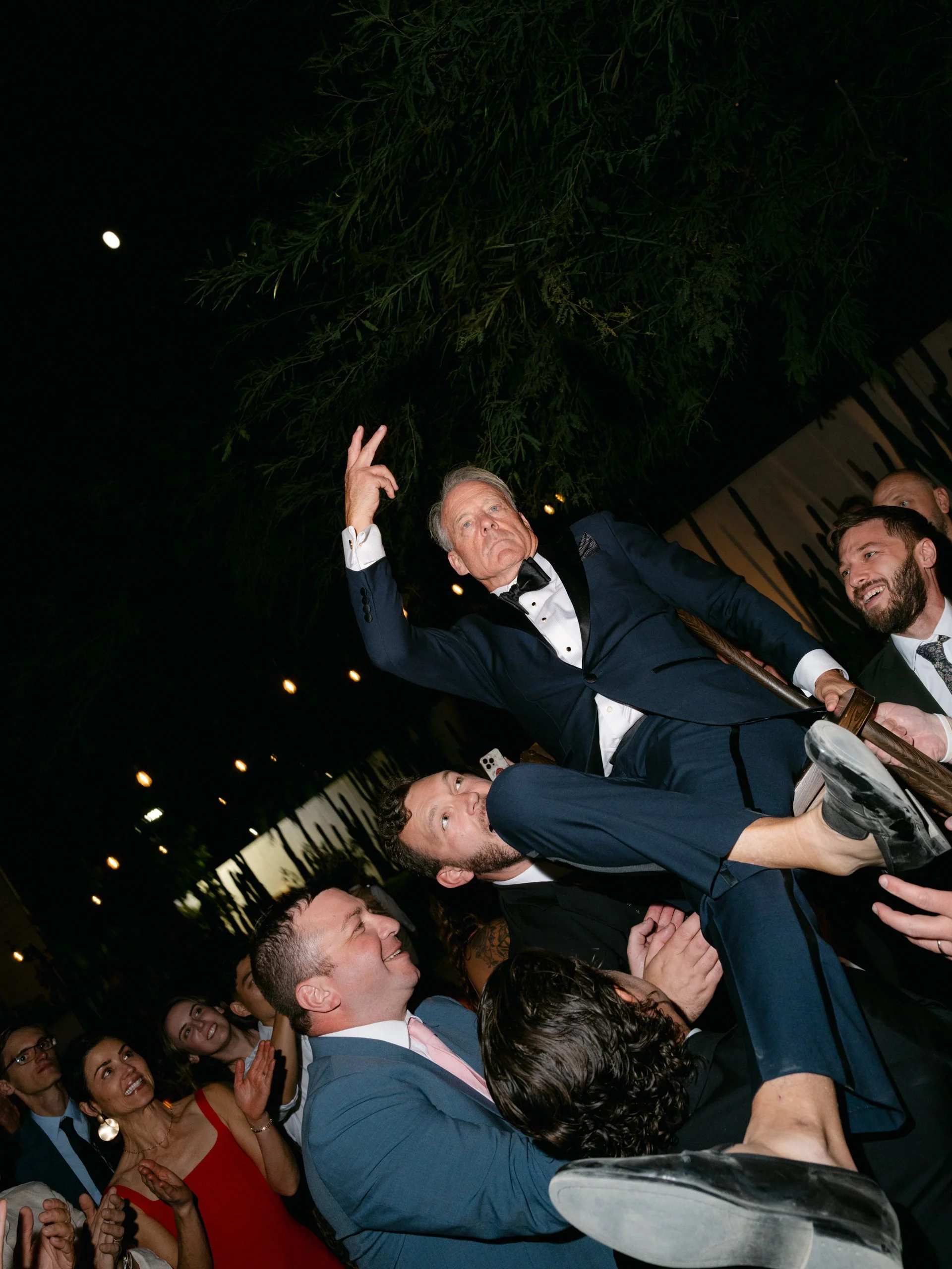 A man in a tuxedo is being lifted and held horizontally by a group of people, while he strikes a confident pose with one hand raised, during a lively celebration at night.