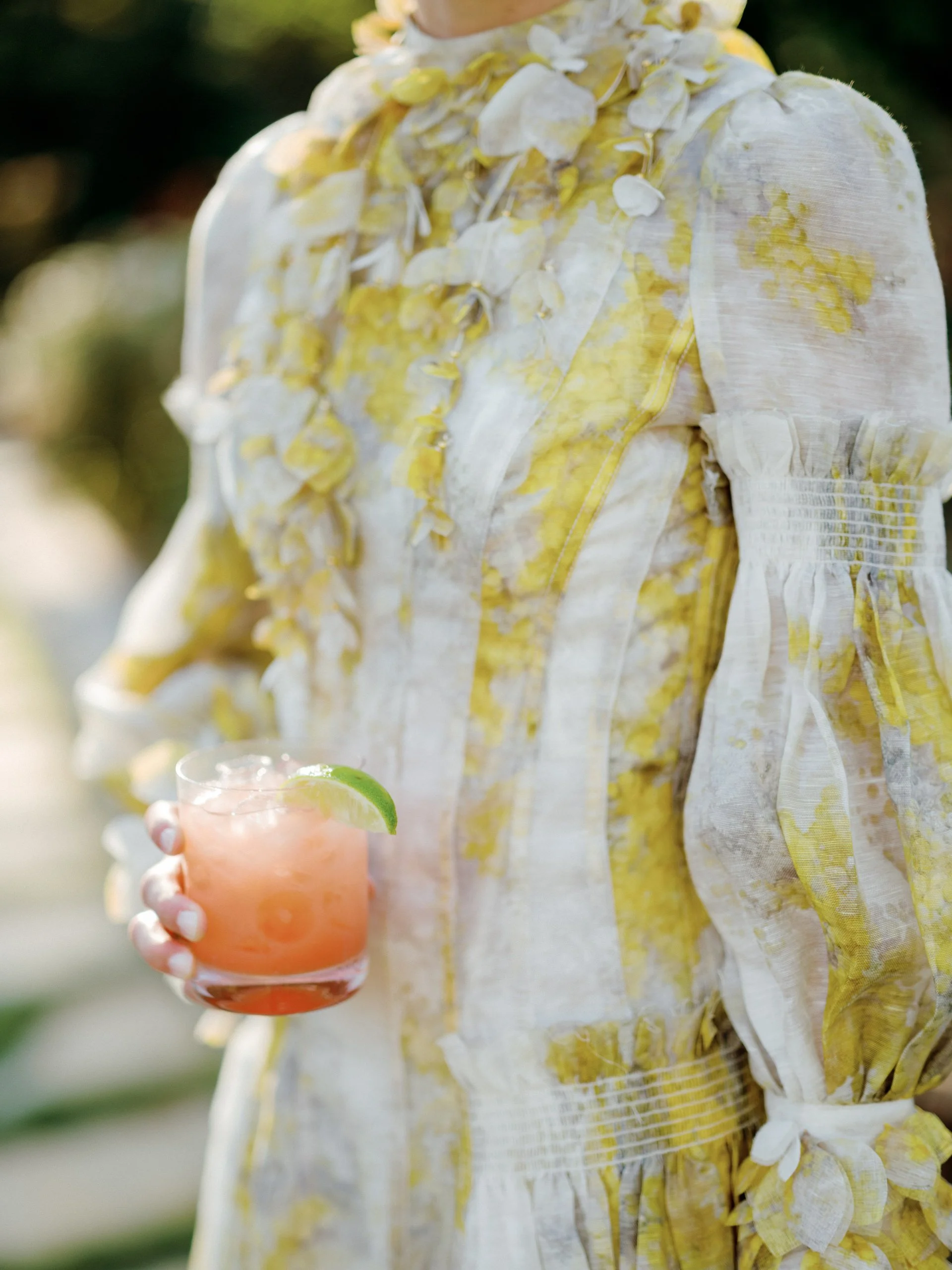 Person holding a pink cocktail with ice and a lime wedge, wearing a white and yellow floral dress.