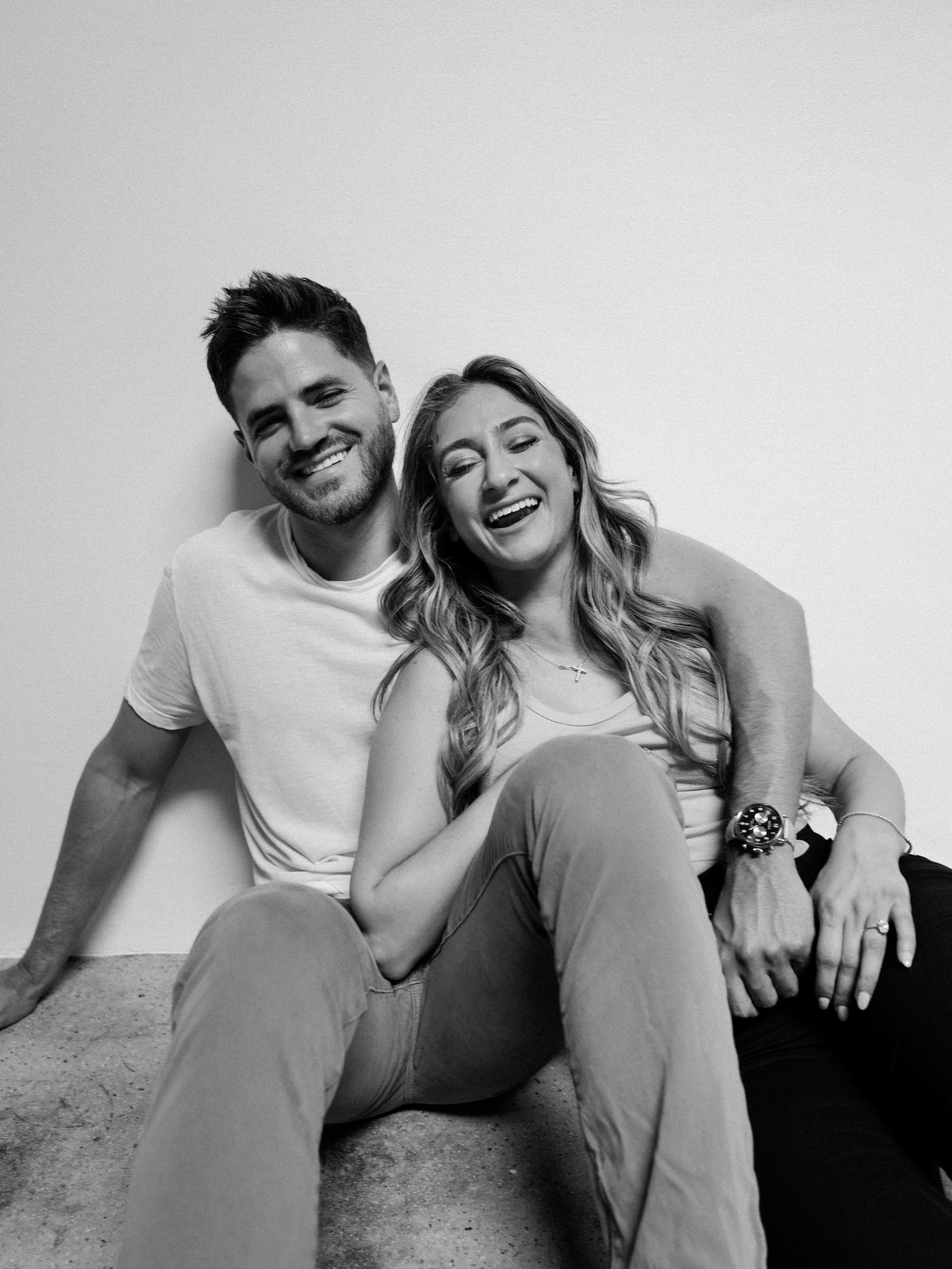 A smiling man and woman sitting on the floor against a plain wall, appearing relaxed and happy.