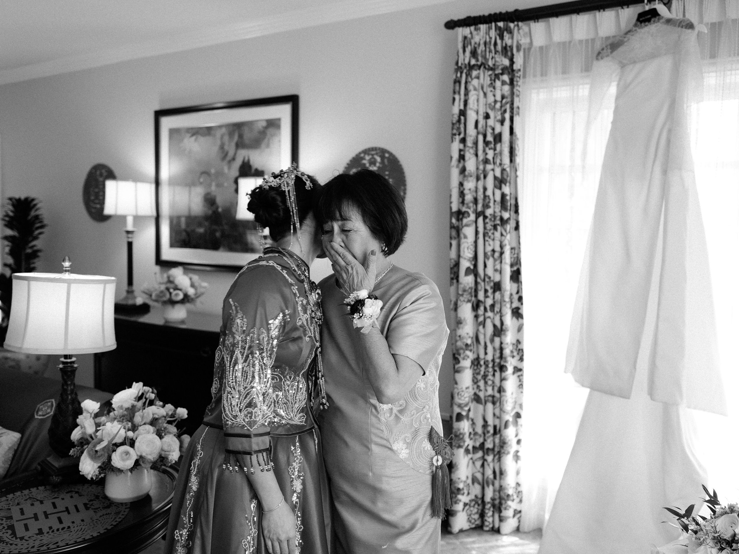 A young woman in traditional Asian attire and an older woman share a tender moment, touching foreheads and laughing inside a warmly lit room with floral curtains and a wedding dress hanging near a window.