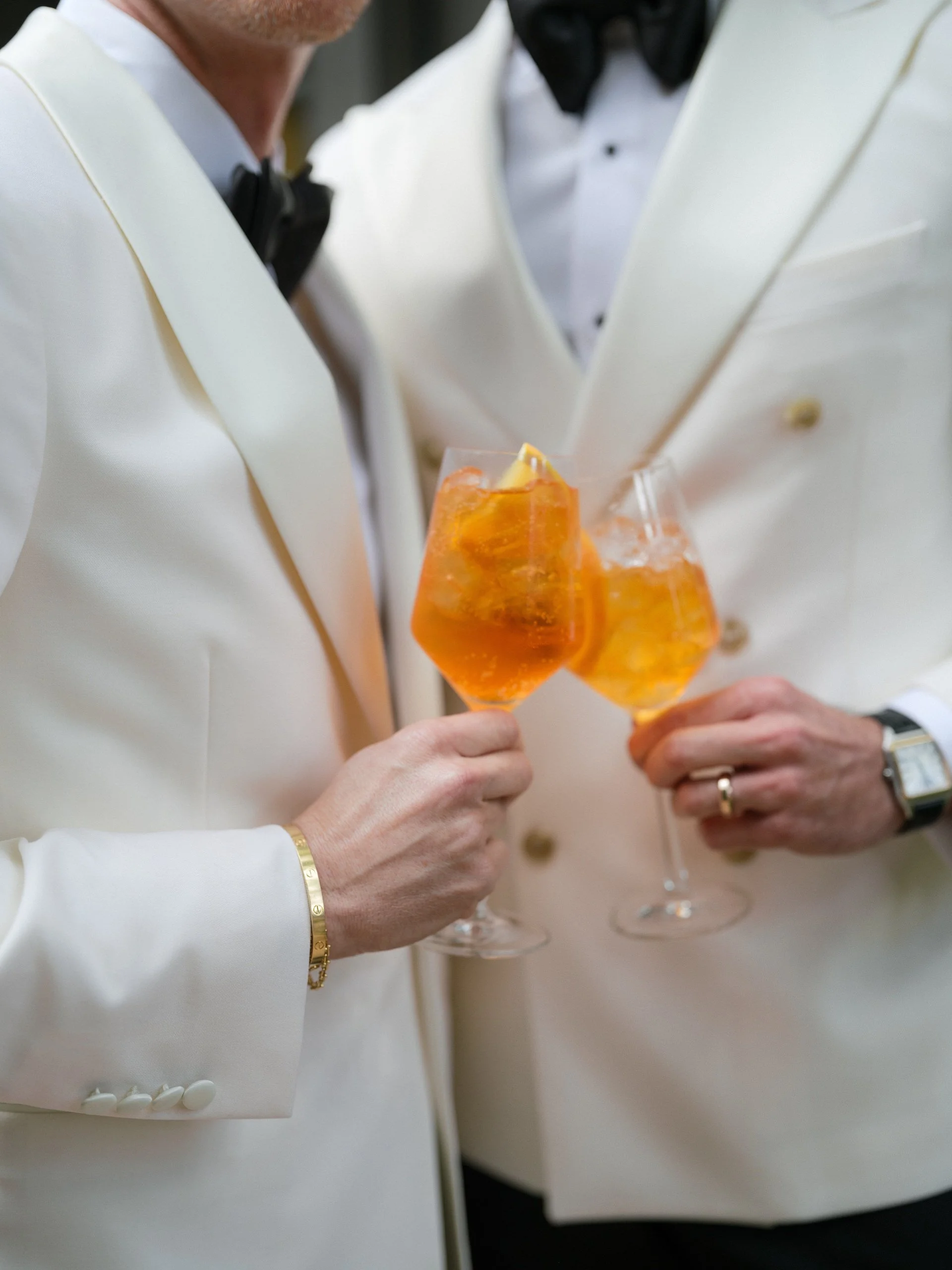 Two people dressed in formal white tuxedos with black bow ties holding champagne glasses with orange-colored drinks and ice.