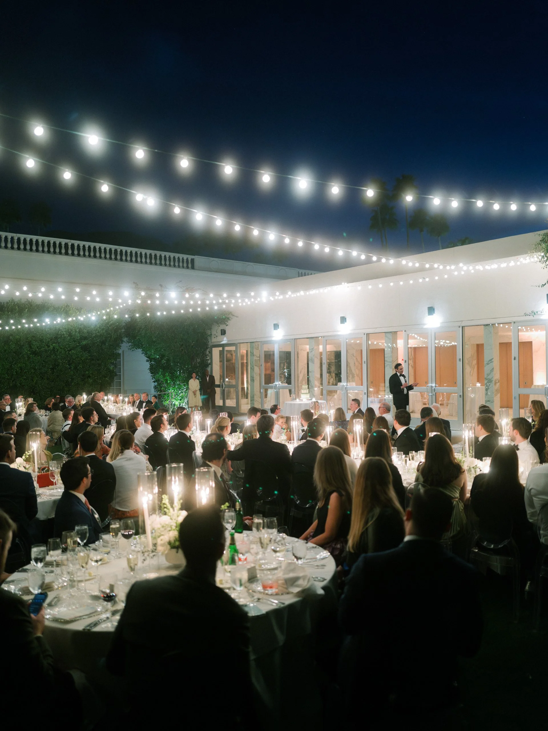 A formal outdoor evening event with many guests seated around round tables, illuminated by string lights overhead, with a man speaking at a podium in front of a building with large glass doors.