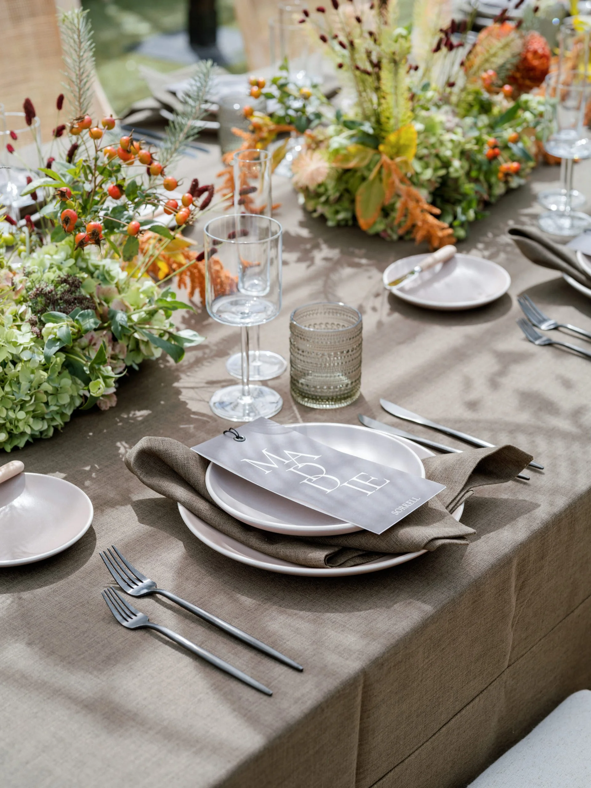 Elegant table setting with white plates, silverware, and a beige napkin, featuring a black and white menu card on top. The table is decorated with lush floral centerpieces and glassware, all set on a beige tablecloth for a formal event.