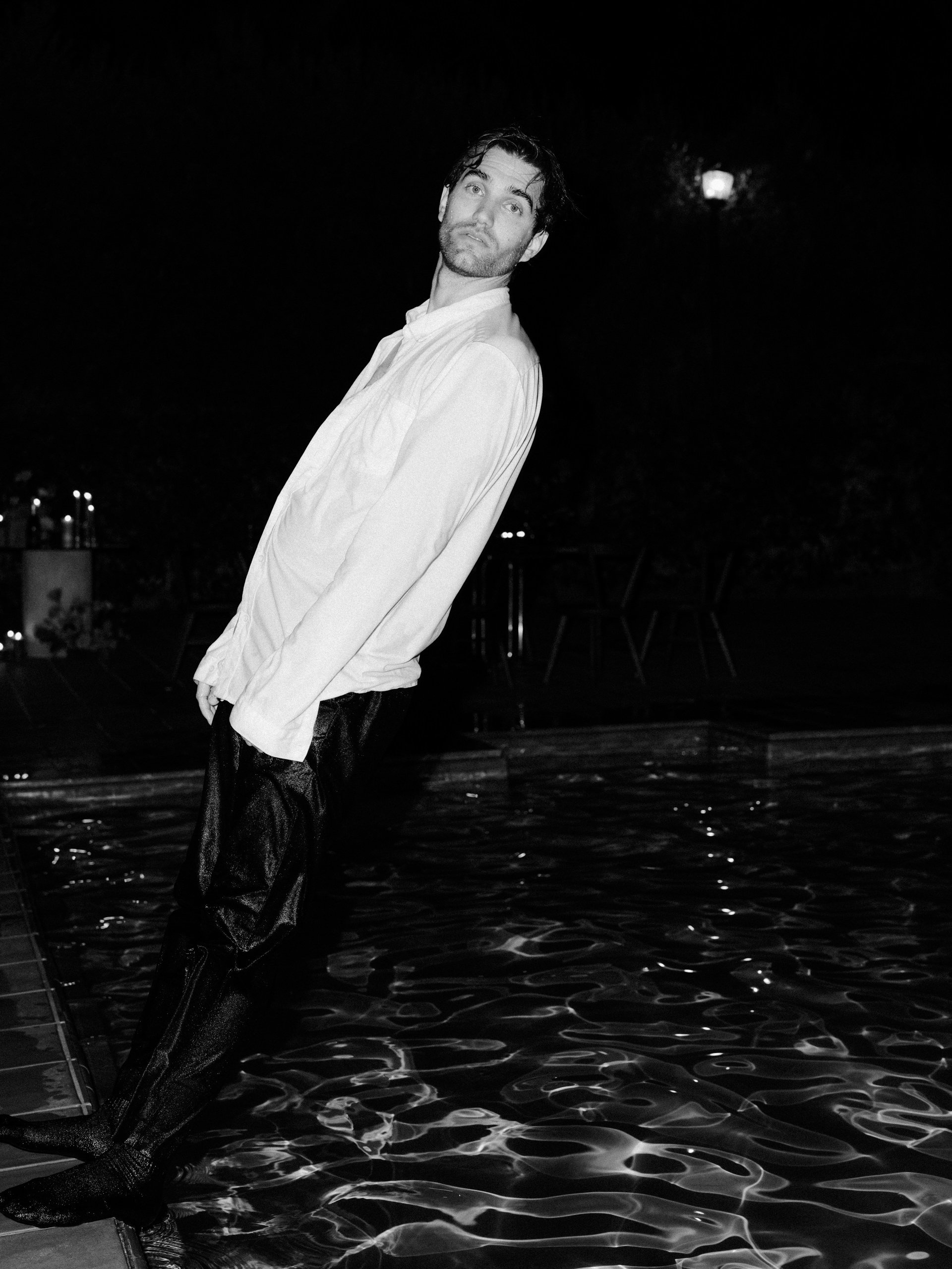 A man with wet hair is standing at the edge of a swimming pool at night, wearing a light-colored shirt and dark pants, with one foot submerged in the water.