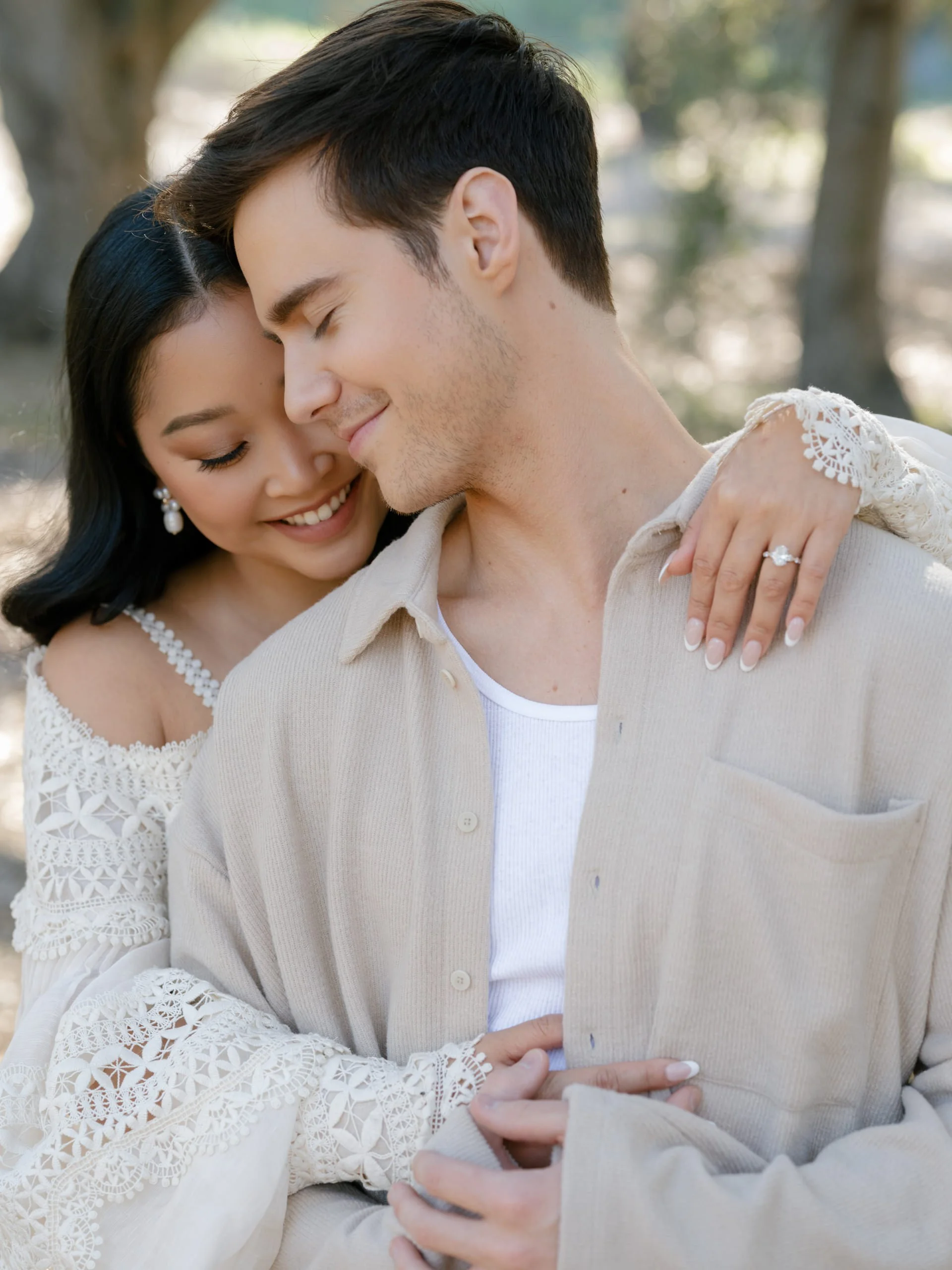 A happy couple with eyes closed, smiling and embracing outdoors, surrounded by trees.