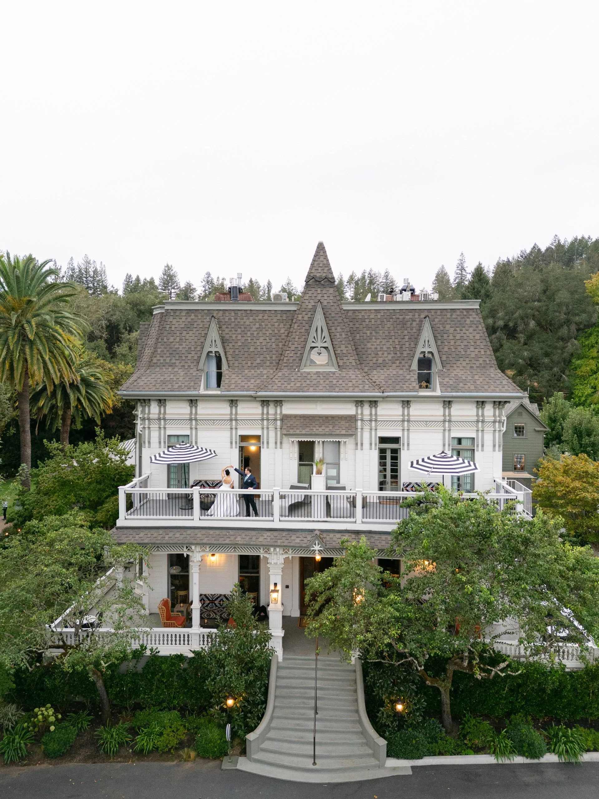 A large, Victorian-style house with a prominent tower, white exterior, and multiple windows is shown surrounded by trees. A couple is standing on the second-floor balcony, likely during a wedding, with umbrellas providing shade.