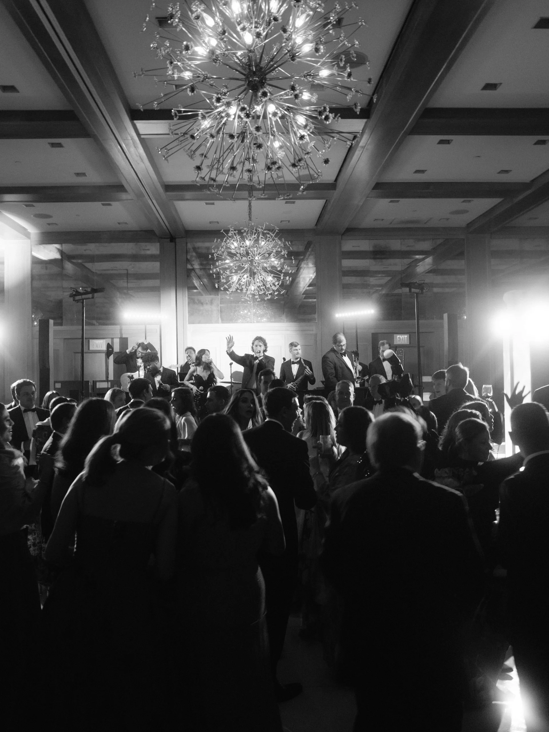 Black and white photo of people dancing and socializing at a formal event or party with a live band playing on stage under large decorative chandeliers.