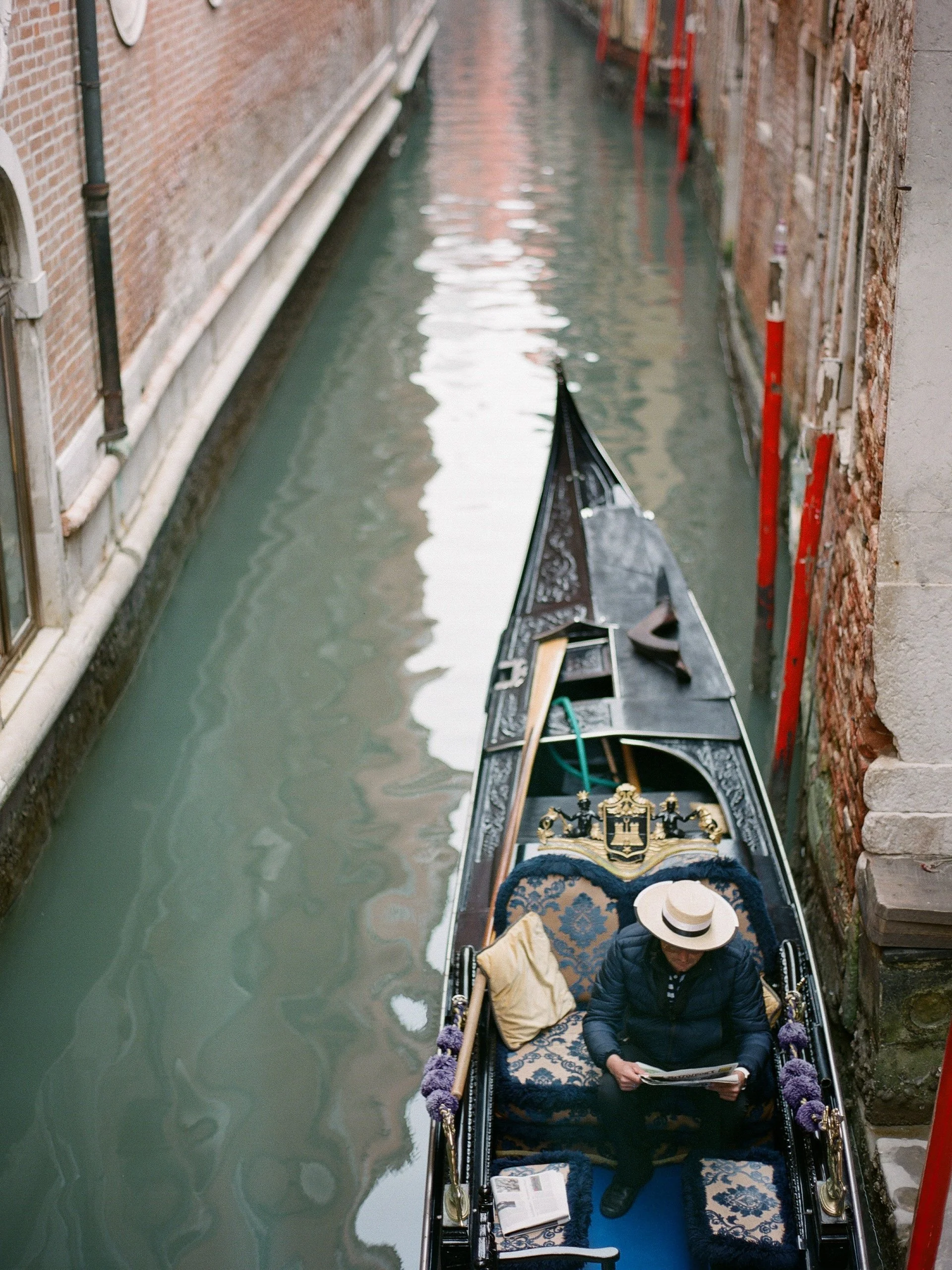 A person wearing a hat and dark clothing sitting in a gondola on a narrow canal, reading a newspaper.