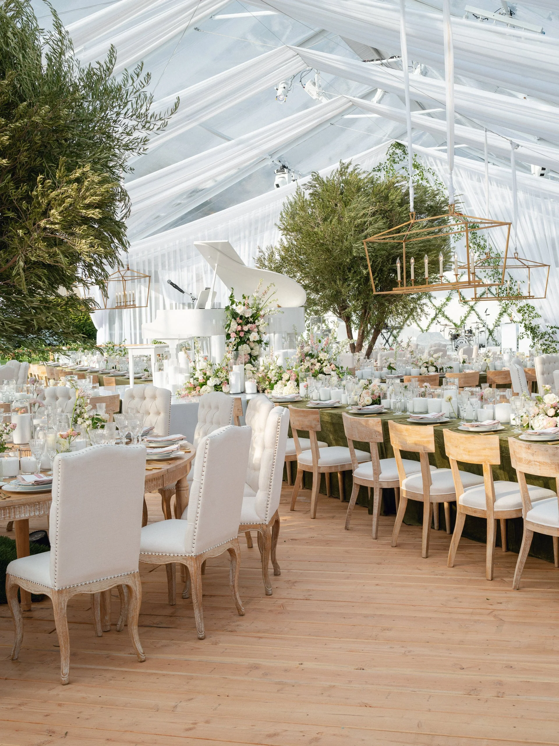 Elegant indoor event space with light wooden floors, decorated with white chairs, floral centerpieces, and greenery, featuring a white grand piano and a chandelier.