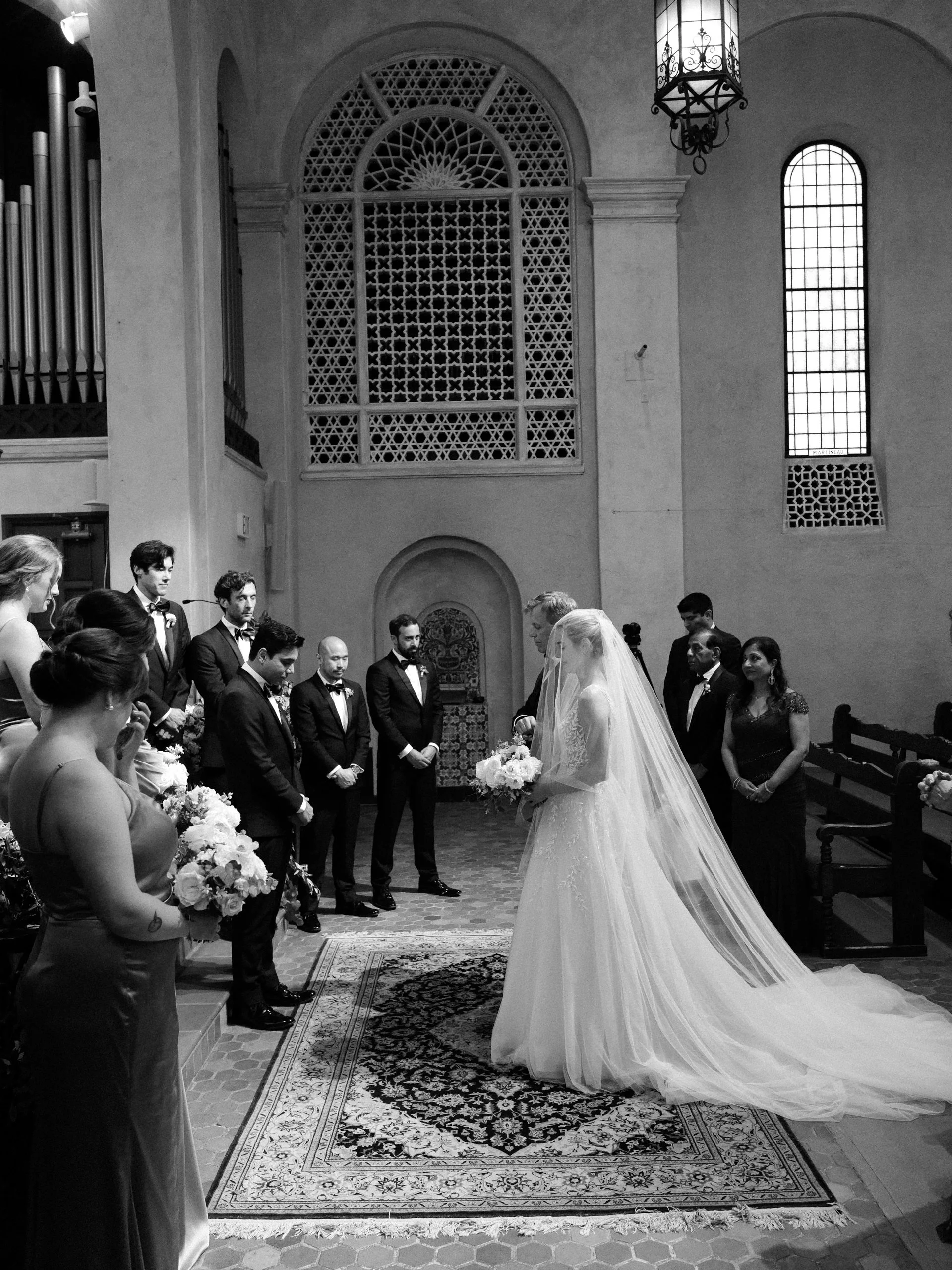 A black and white photograph of a wedding ceremony inside a church with high ceilings and tall windows. The bride, wearing a long veil and holding a bouquet, is standing at the altar with her wedding gown flowing behind her. The groom and groomsmen, 