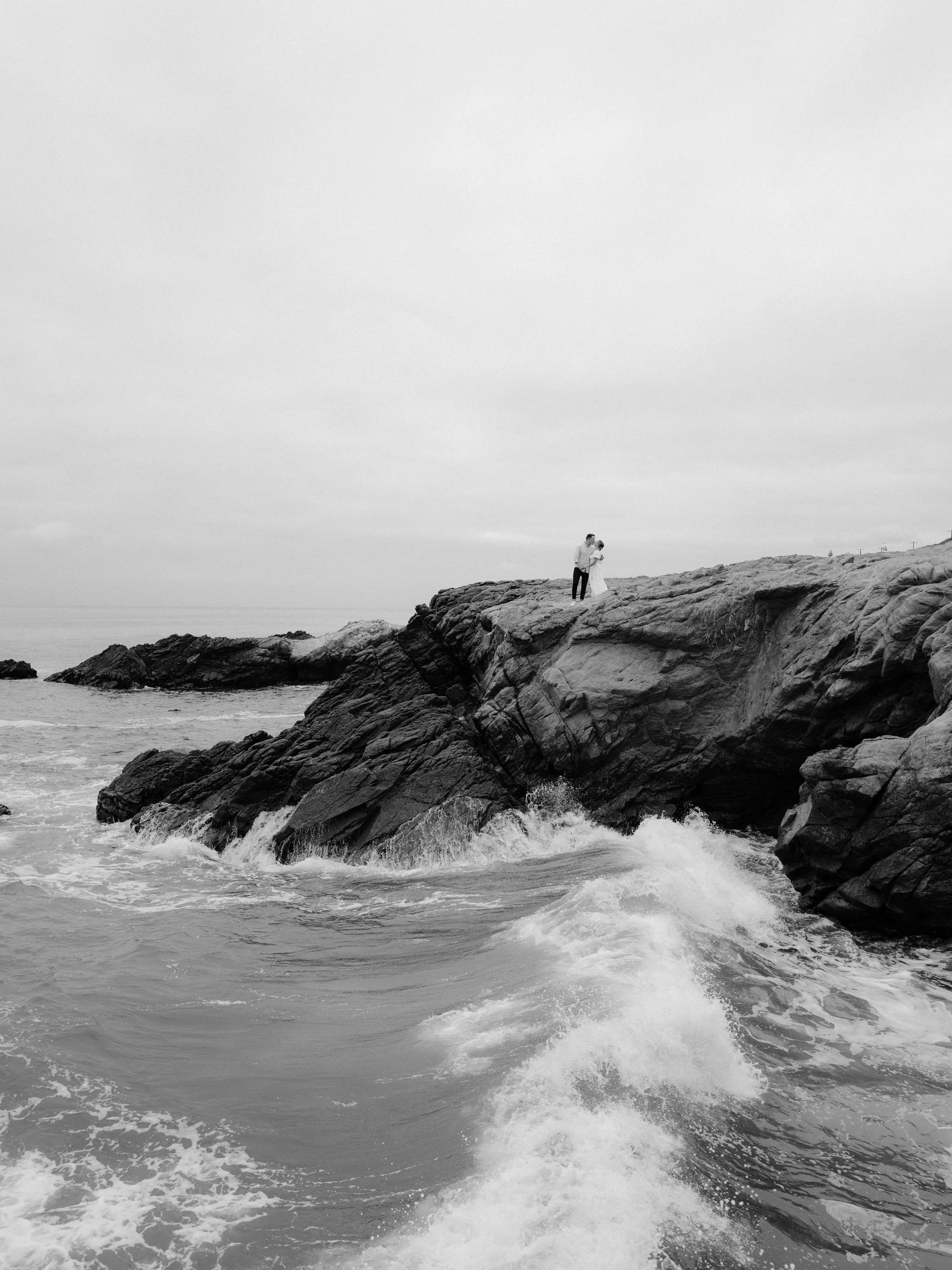A couple standing on a rocky outcrop by the ocean, holding each other, in a black-and-white photo.