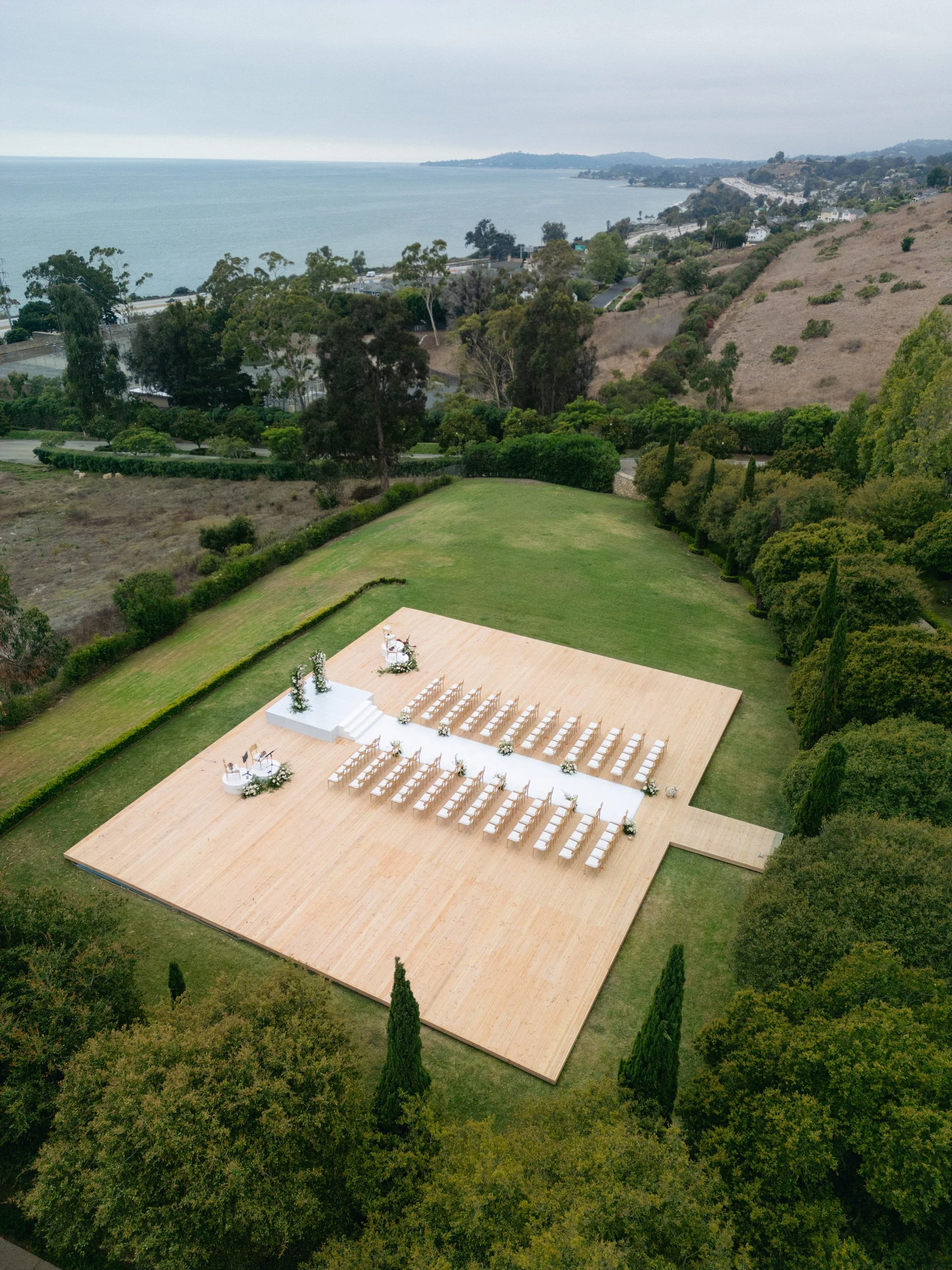 Outdoor wedding ceremony setup on a wooden platform with rows of white chairs, floral decorations, and a view of a coastline with trees and hilly terrain in the background.
