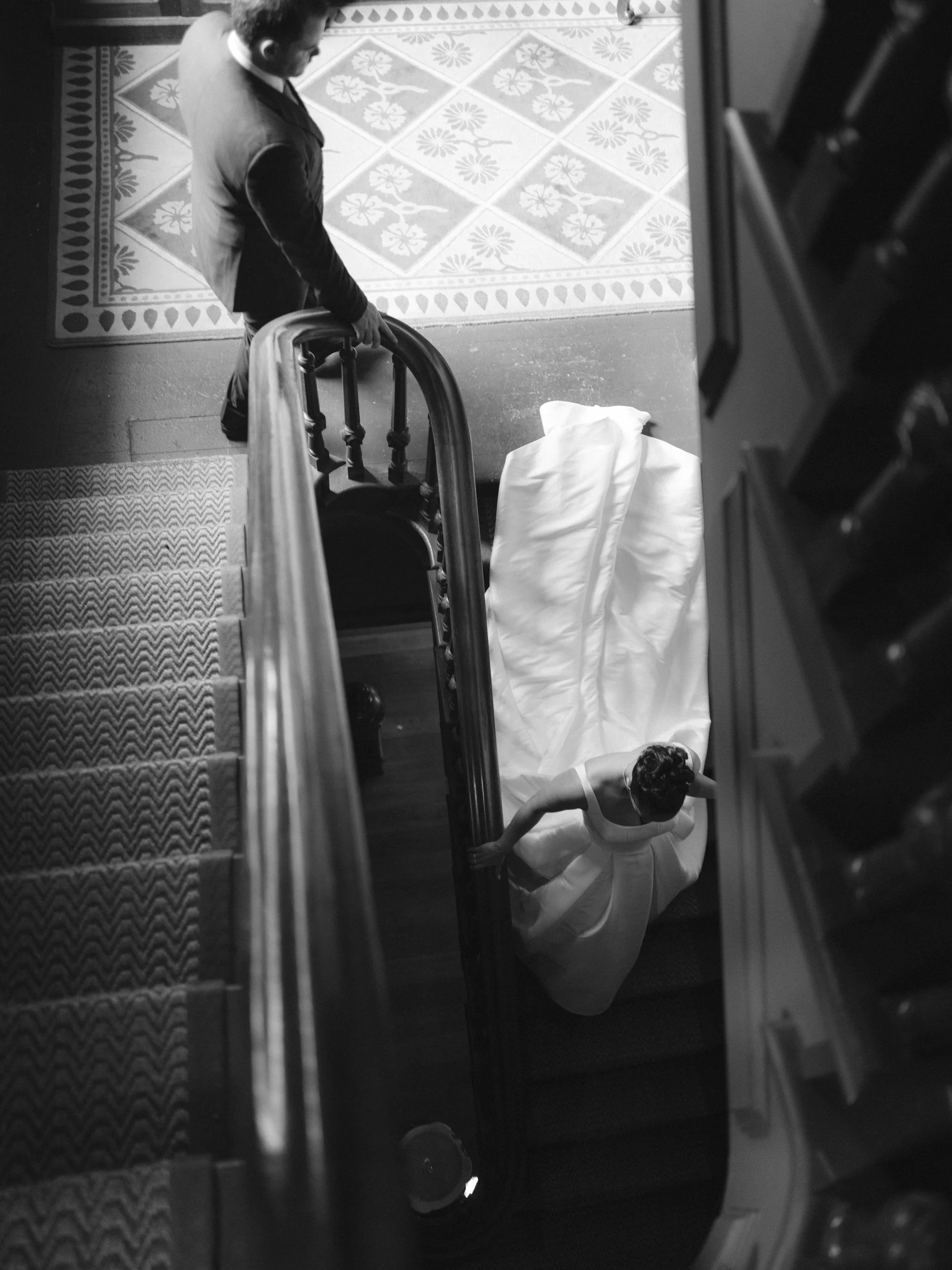 A bride in a wedding dress standing on stairs, viewed from above, with a man at the top of the stairs looking down at her.