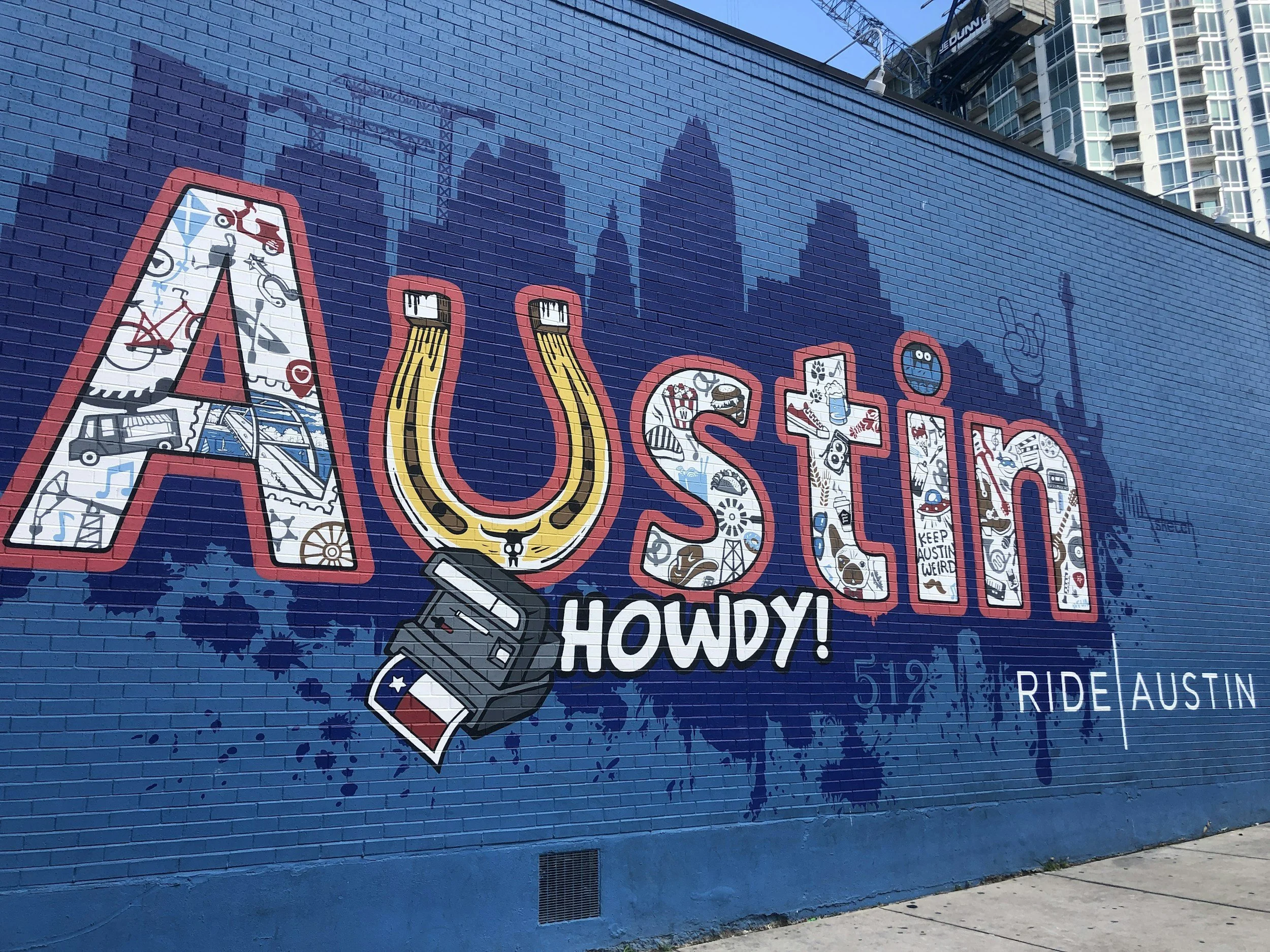 Colorful mural on a blue brick wall with the word 'AUSTIN' decorated with various illustrations inside the letters, and the words 'HOWDY!' and 'RIDE AUSTIN' below.