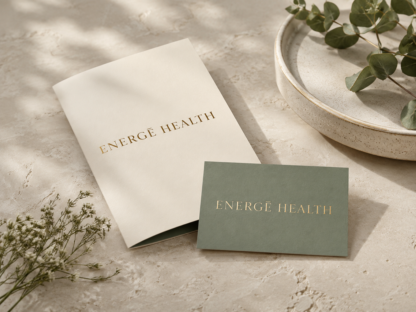 A booklet and a card with the words 'ENERGĒ HEALTH' on them placed on a light-colored surface with a bowl containing green leaves nearby.