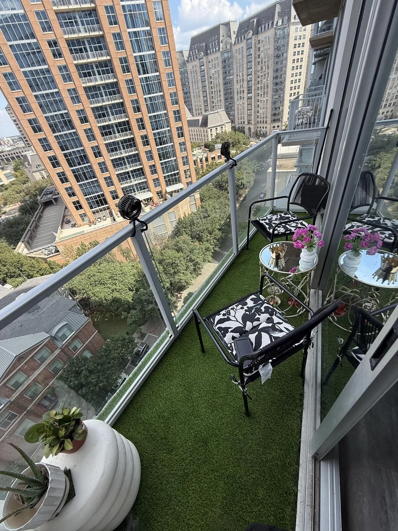 A small balcony with artificial grass flooring, black chairs with floral cushions, and a glass-topped side table with pink flowers in a white vase, overlooking tall city buildings and a park below.