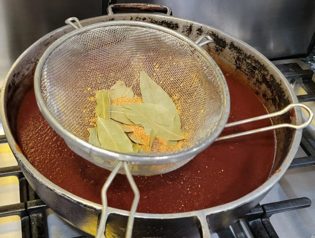 A skillet of simmering tomato sauce with bay leaves and spices, with a metal strainer holding more bay leaves above it.