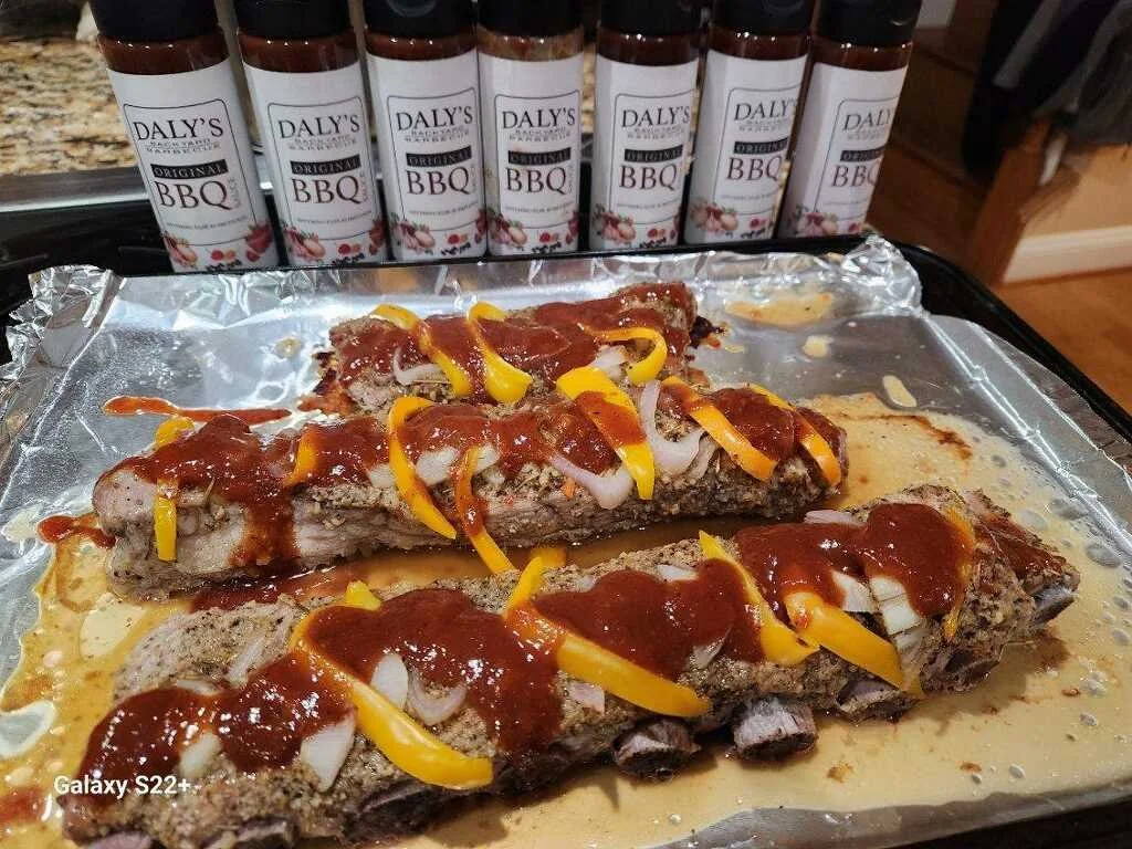 Two cooked meatloaves topped with ketchup, sliced onions, and yellow bell peppers, with multiple bottles of Daly's Original BBQ sauce in the background, on a baking sheet covered with aluminum foil.
