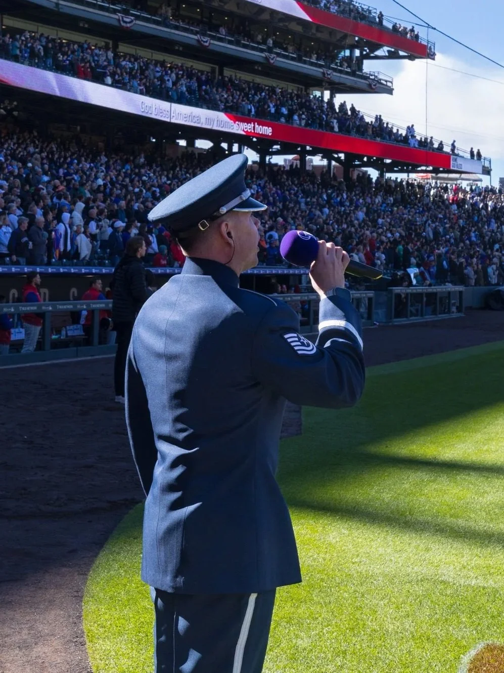 A uniformed military officer with a cap, holding a microphone, stands on a baseball field in front of a packed stadium during a patriotic event at a baseball game.