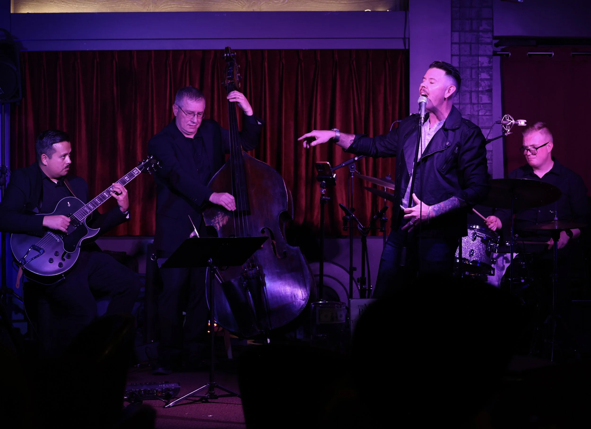A band performing on stage with four musicians, one singing into a microphone, two playing guitars, one on bass, and one on drums, against a backdrop of red curtains with purple stage lighting.