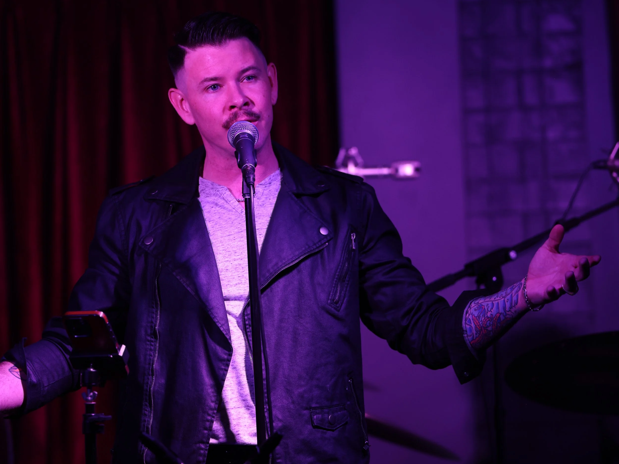 A man with dark hair, styled in a pompadour, is standing on stage in front of a microphone, wearing a black leather jacket and a gray shirt. The stage has purple lighting, red curtains, and some musical equipment in the background.