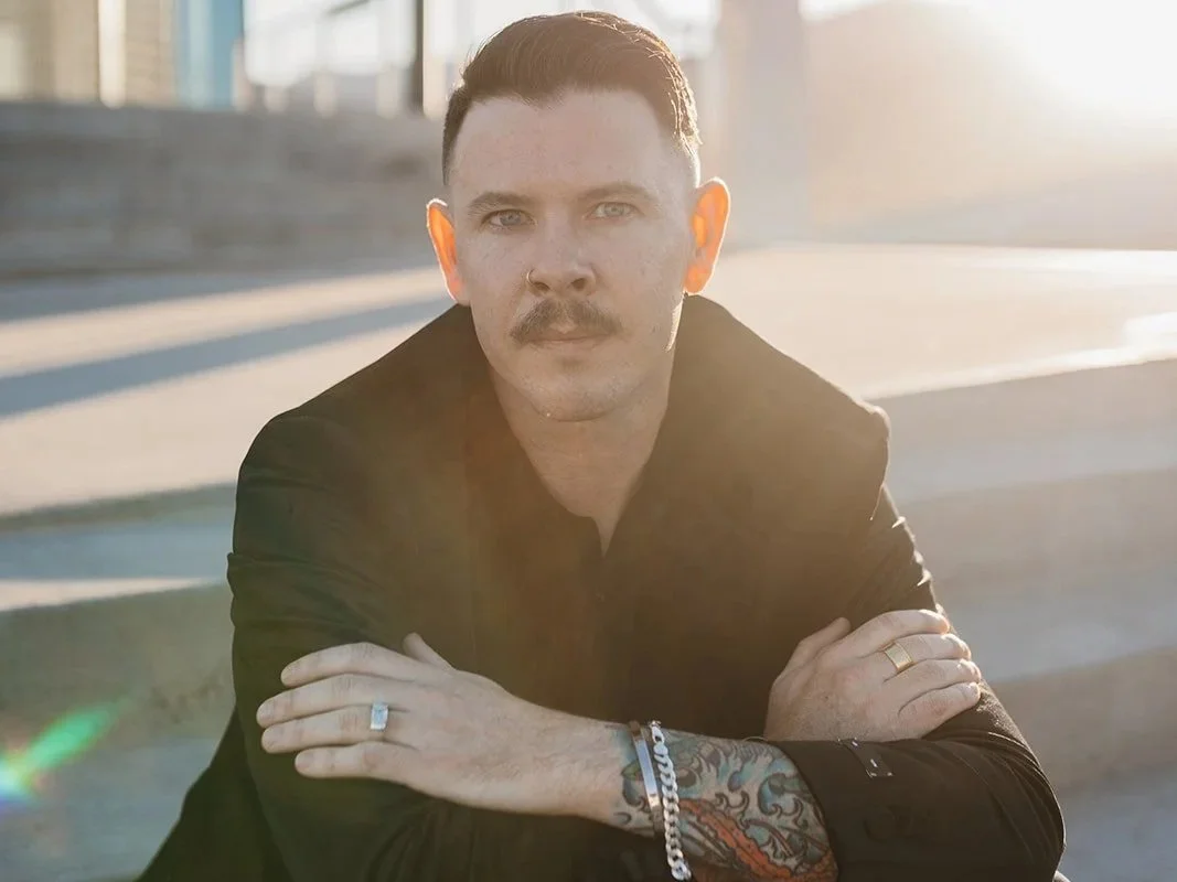 A man with a moustache and tattoos on his arm, wearing a black jacket, is sitting outdoors with crossed arms, illuminated by sunlight.