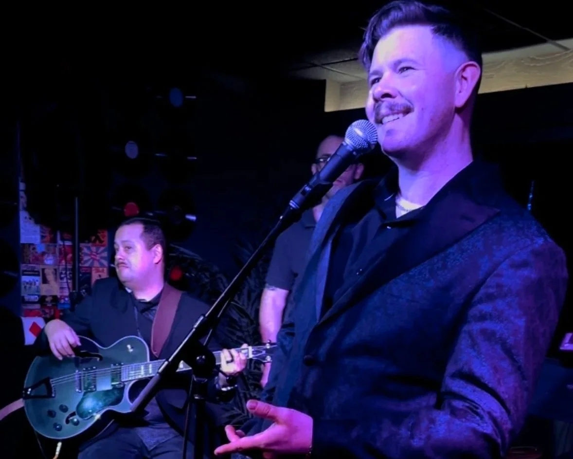 A man with a mustache and styled hair singing into a microphone, standing in front of two men playing guitars on a stage with dark background and posters on the wall.