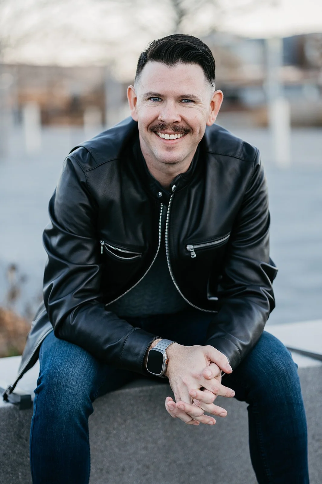 A smiling man with short dark hair and a mustache, wearing a black leather jacket, dark jeans, and a smartwatch, sitting outdoors on a concrete surface, with a blurred background of trees and buildings.