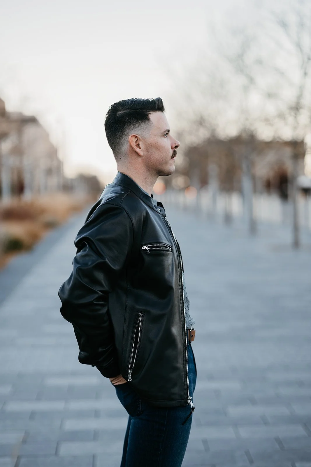Side profile of a man wearing a black leather jacket standing outdoors on a pathway with trees and buildings in the background.