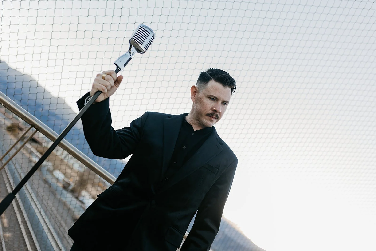 A man in a black suit holding a vintage microphone, standing outdoors near a wire mesh fence with mountainous scenery in the background.