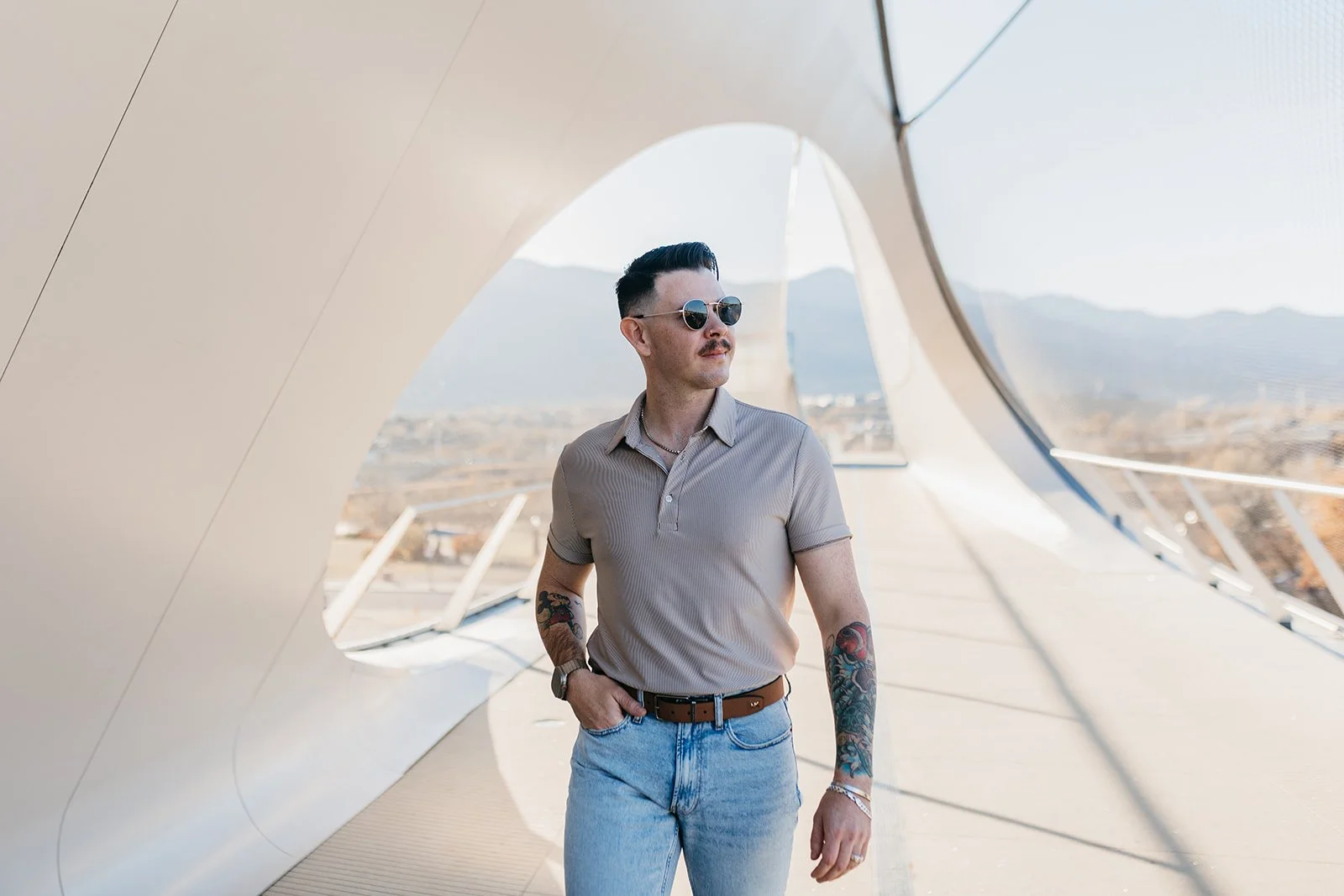 A man with sunglasses, a mustache, and tattoos on his arms stands under a modern architectural structure outdoors, with mountains in the background.