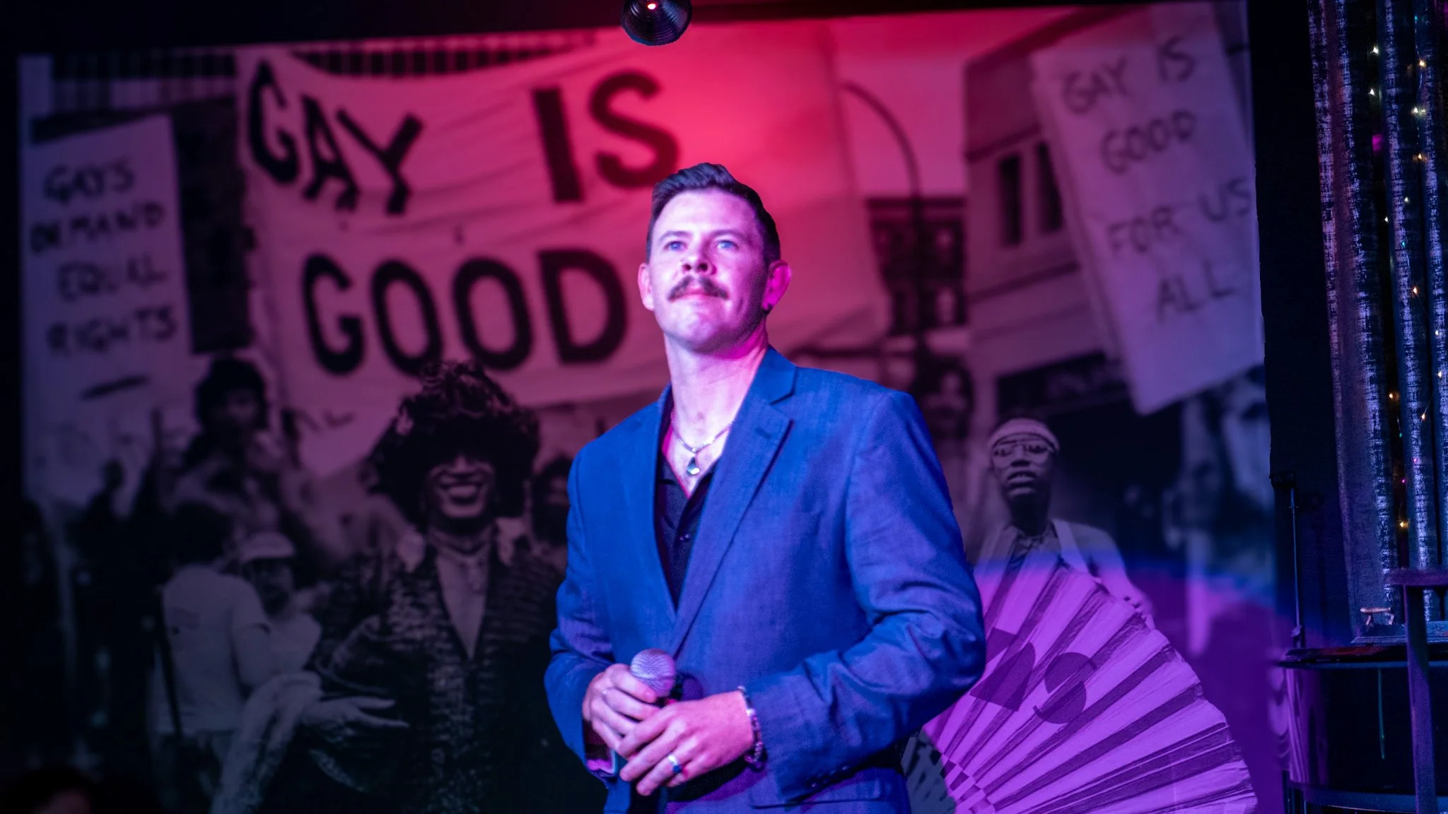 A person with a mustache in a blue suit holding a microphone on stage with a colorful background that includes signs saying "GAY IS GOOD" and "GAY IS GOOD FOR US ALL" and images of diverse people.