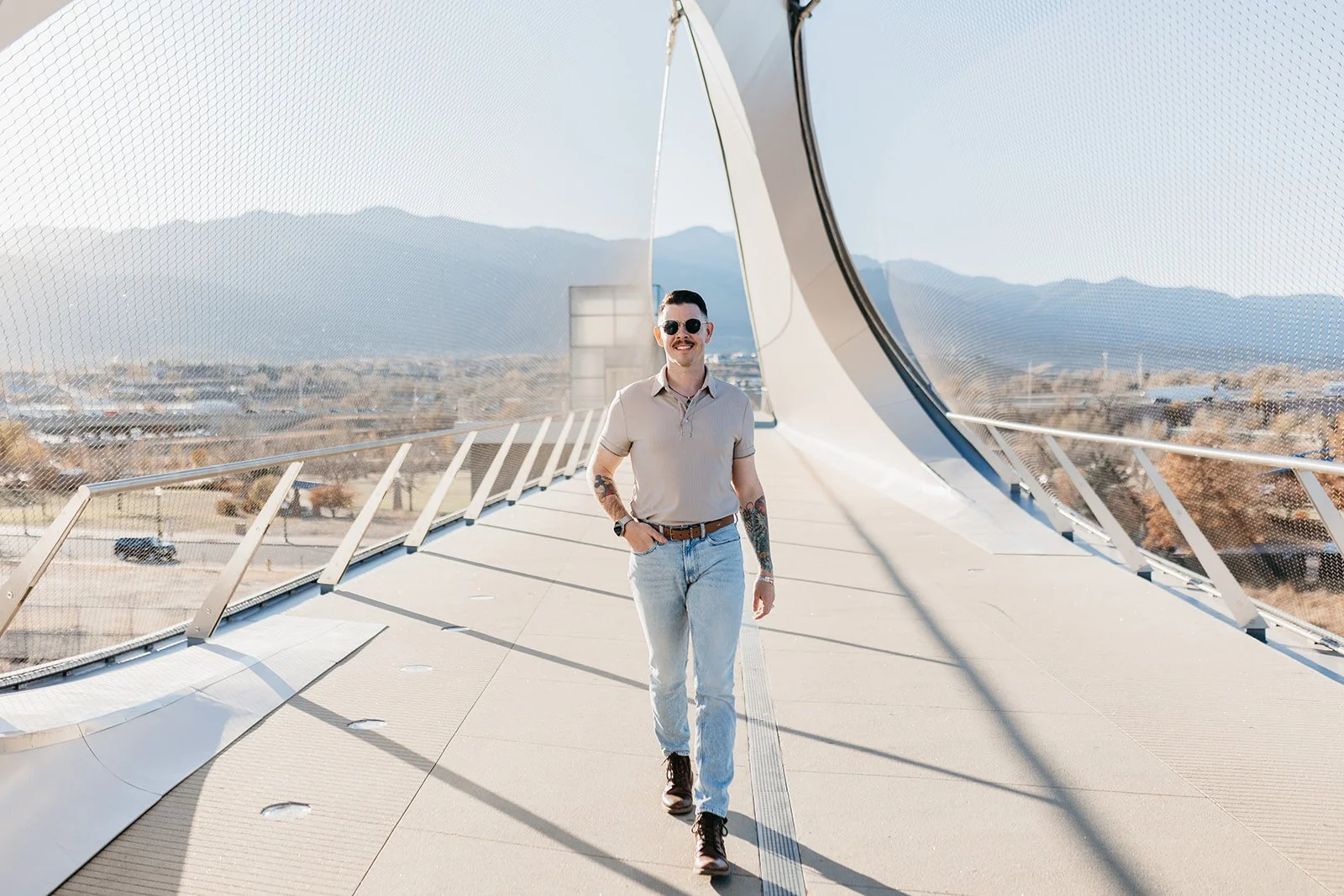 A man walking on a modern outdoor bridge with a mountain view in the background. He is wearing sunglasses, a beige polo shirt, light blue jeans, and boots.