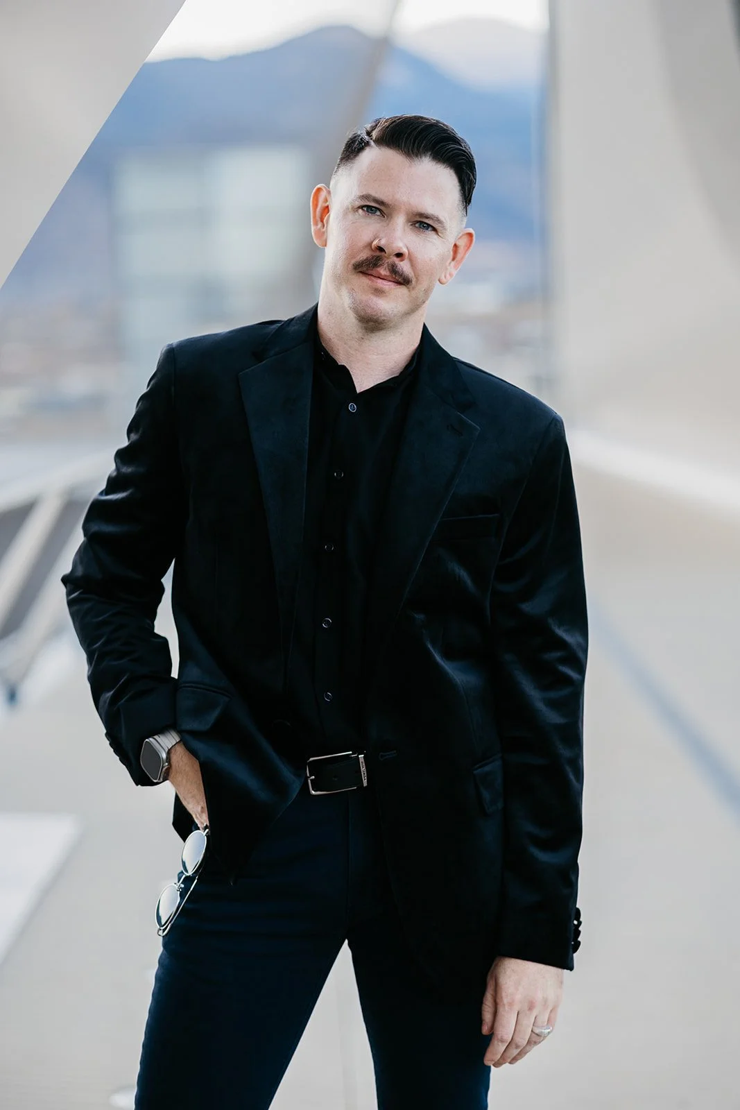 A man with dark hair and a mustache, wearing a black suit and shirt, standing indoors with a modern architectural background and mountains visible through a window.