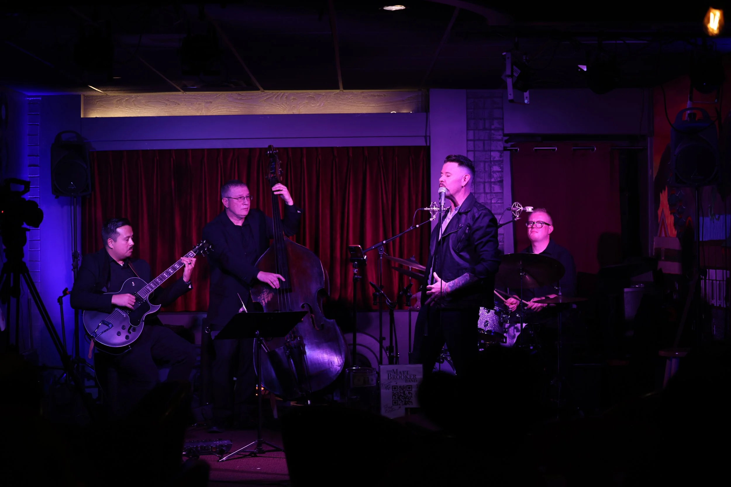 A four-piece jazz band performing on a small stage with red curtains, including a guitarist, a double bassist, a vocalist, and a drummer, under purple stage lighting.