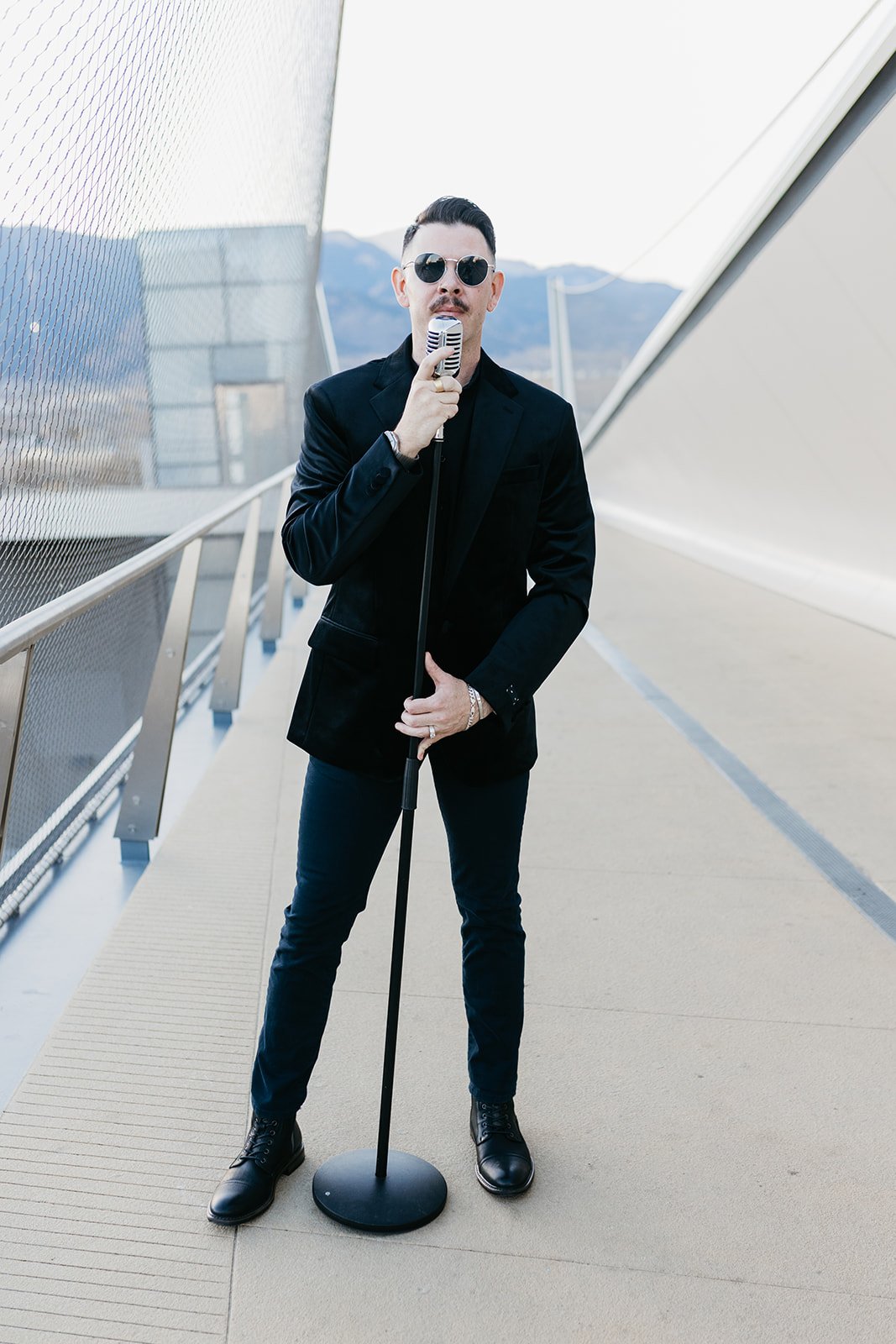 A man in a black suit, sunglasses, and shiny black shoes stands on a modern outdoor walkway, holding a vintage microphone, with a mountain landscape in the background.