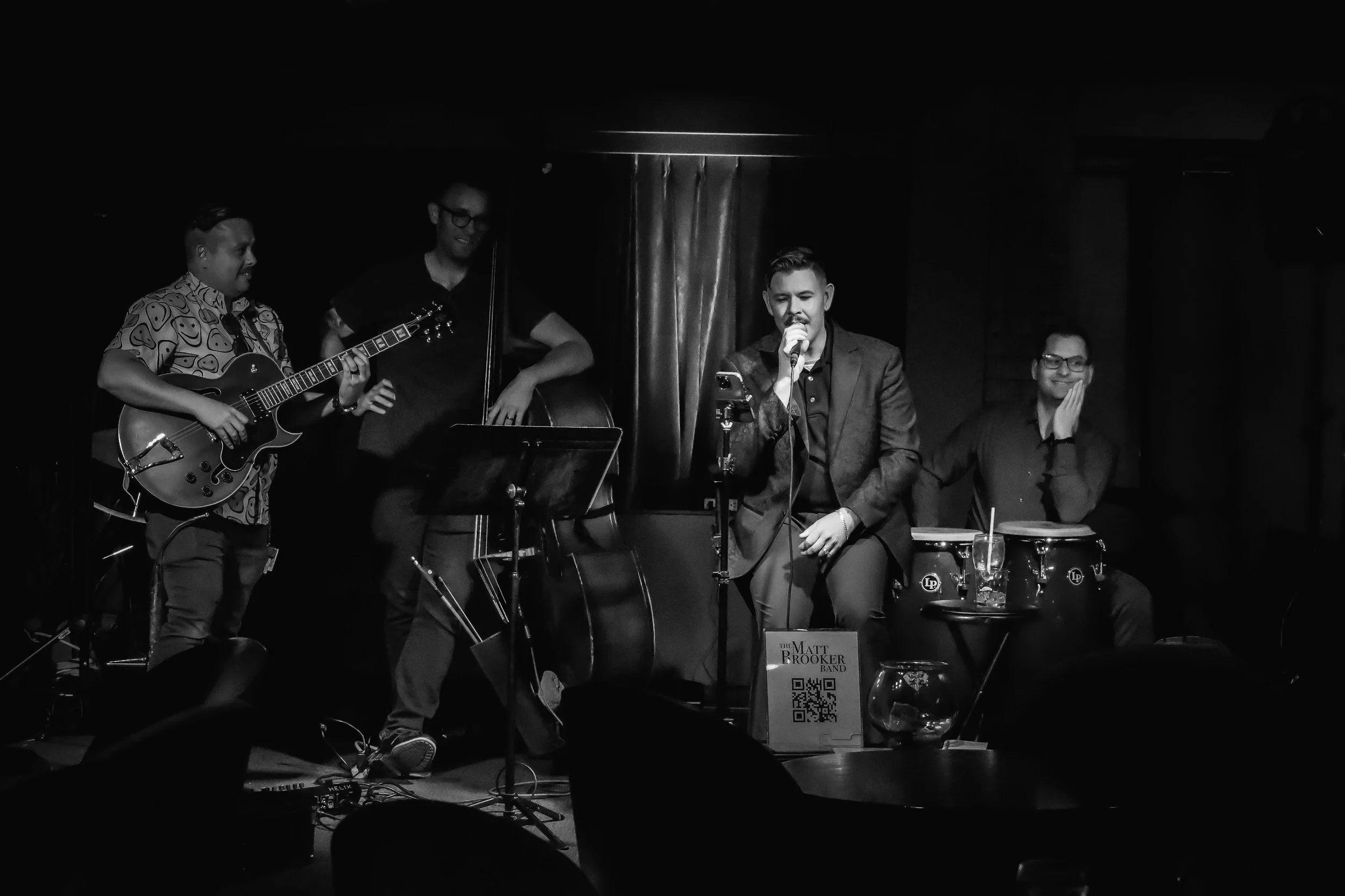A black and white photo of a live jazz band performance with four men. One is singing into a microphone while the other three play musical instruments: guitar, double bass, and percussion. The singer is seated, wearing a suit, and the others are standing or sitting, smiling and engaging with the music. There is a sign with a QR code and text reading 'The Matt Brooker Band' in front of the singer.