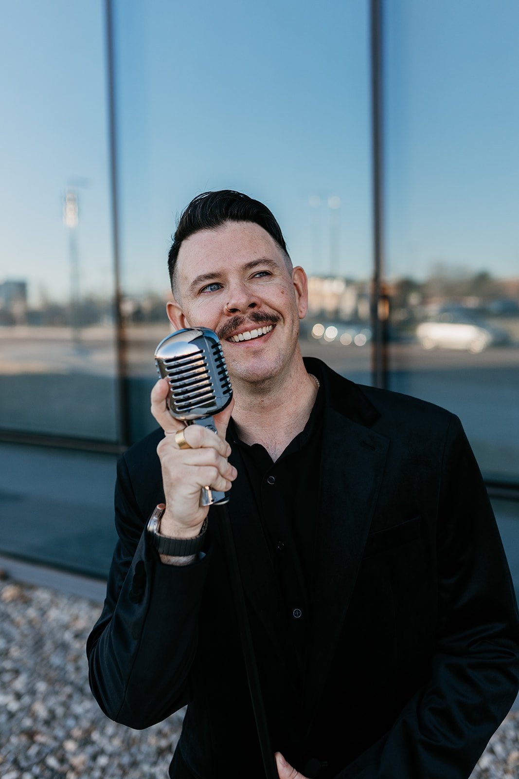 Man with dark hair, mustache, and nose ring singing into a vintage microphone outdoors, wearing a black jacket and shirt, with a reflective glass building in the background.