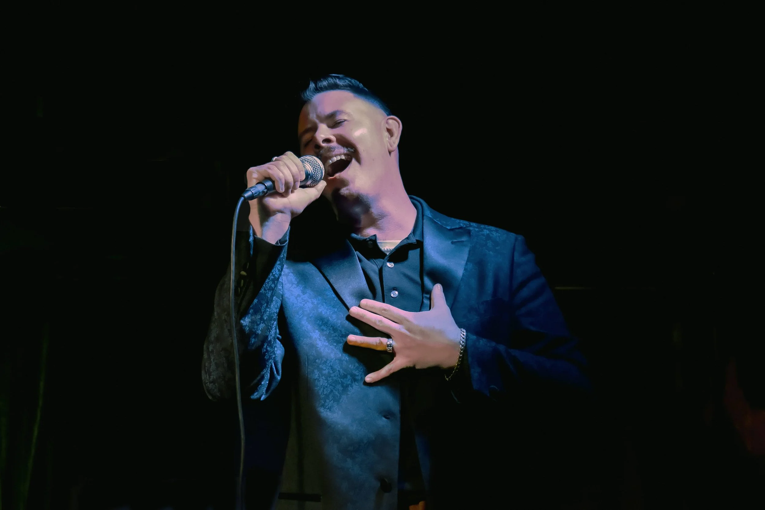 A man singing passionately with a microphone, dressed in a blue suit and black shirt, holding his chest with one hand against a dark background.