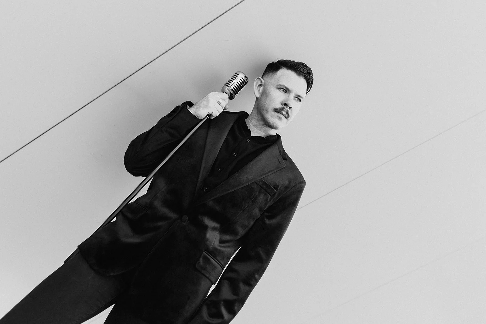 Black and white photo of a man with a mustache and slicked-back hair, wearing a dark suit, holding a vintage microphone, standing against a plain background.