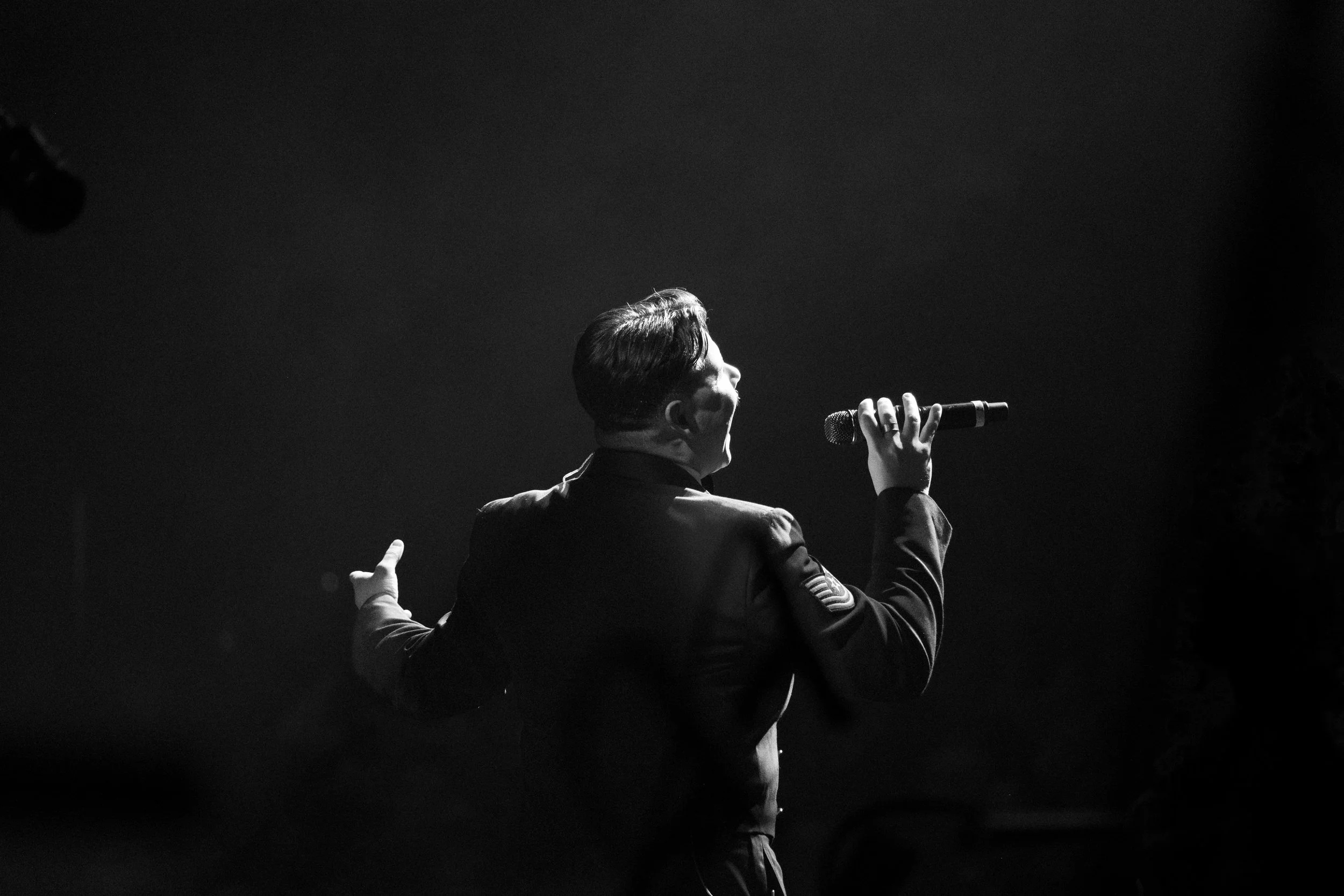 A person, likely a singer, holding a microphone and performing on stage with a dark background.