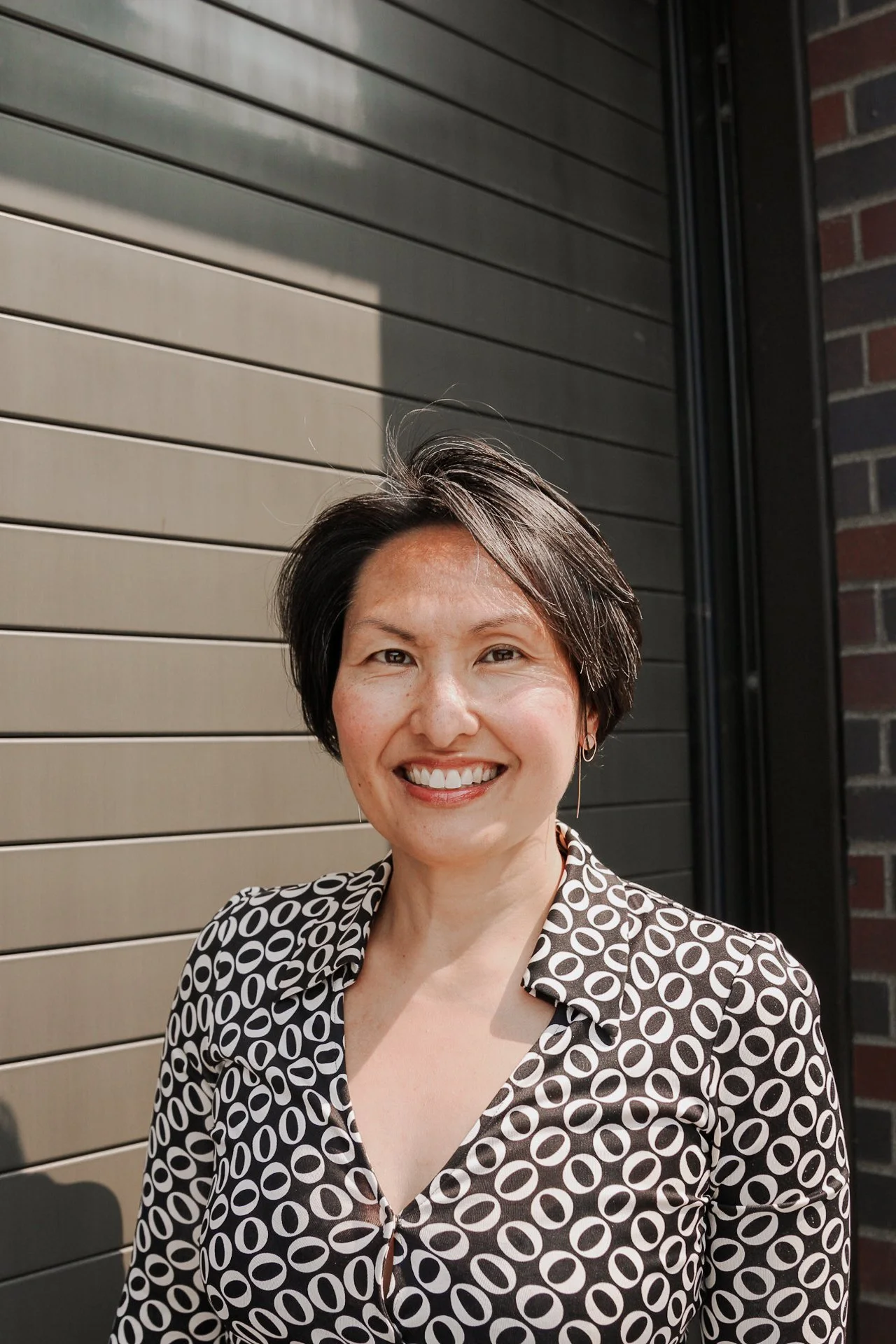 A woman with short dark hair smiling outdoors in front of a metal wall and brick wall, wearing a black and white patterned blouse.