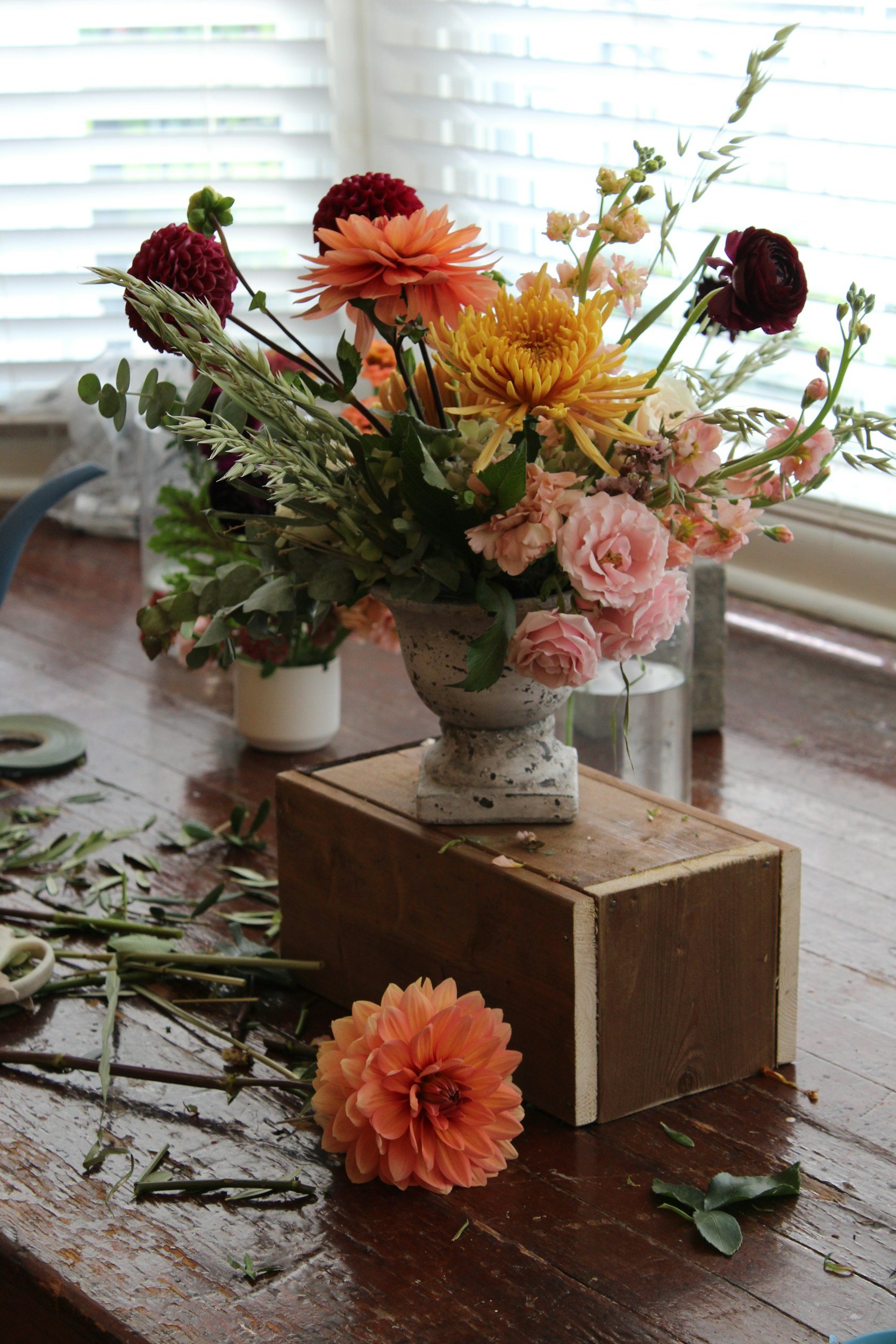 A large floral arrangement in a white vase on a wooden table, with scattered flower stems and leaves around it. The flowers include pink, orange, yellow, and deep red blooms.