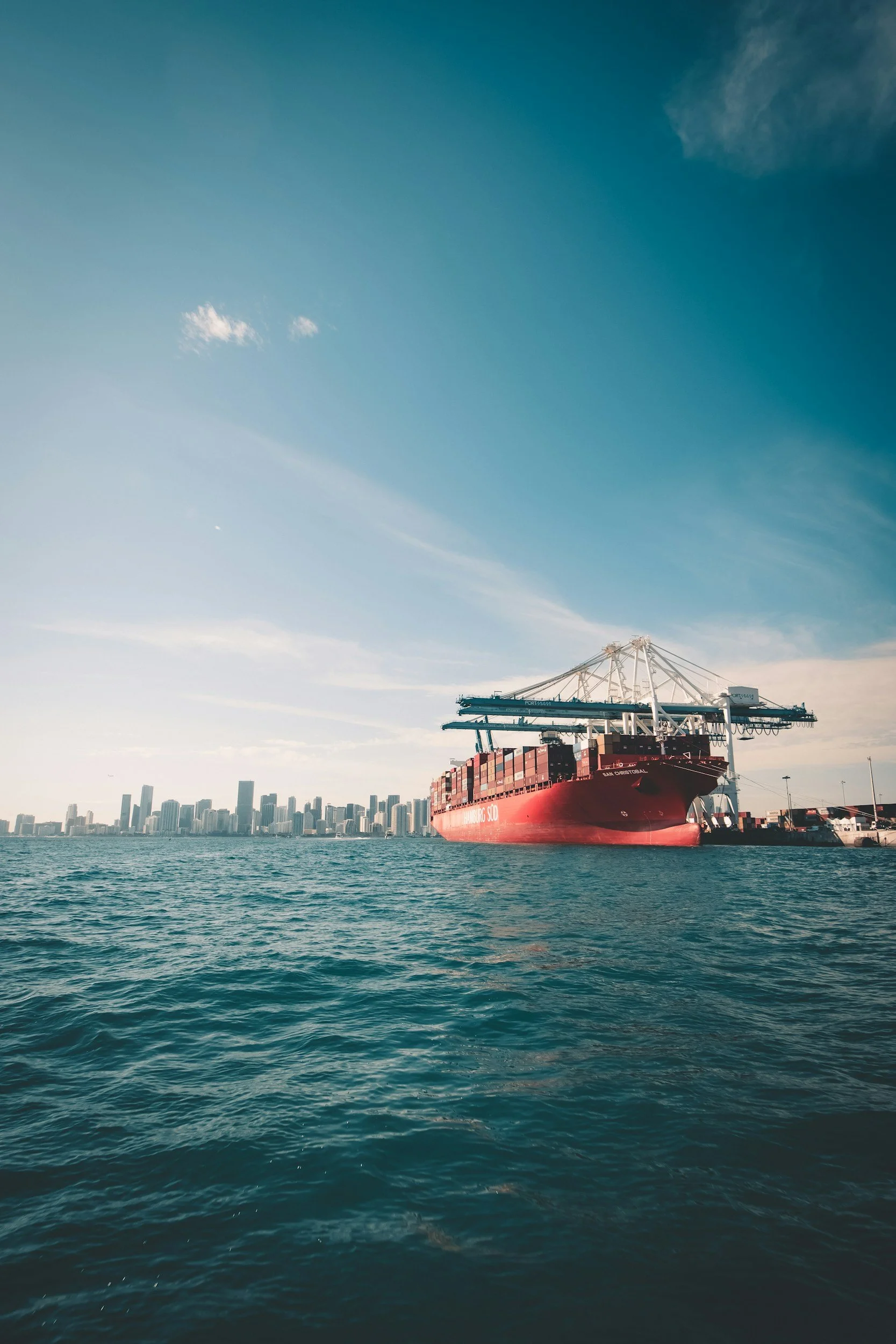 A large cargo ship docked at a port with a city skyline in the background under a partly cloudy sky.