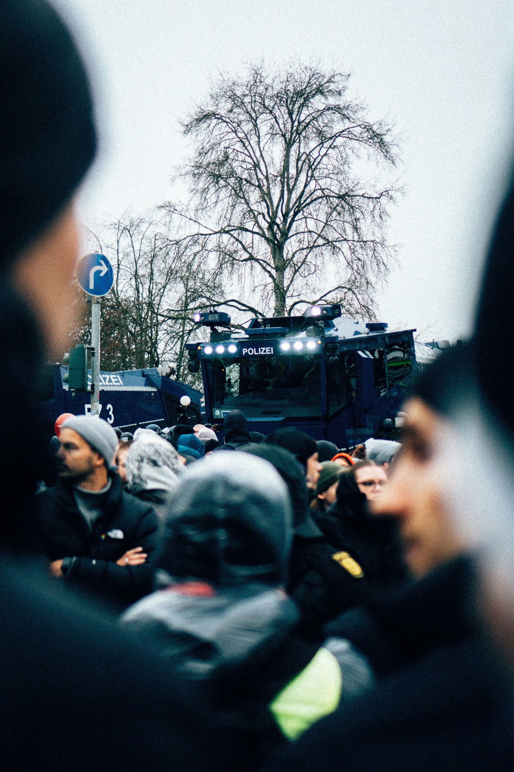 Crowd of people gathered outdoors during daytime, with police vehicles and officers present, and a large leafless tree in the background.