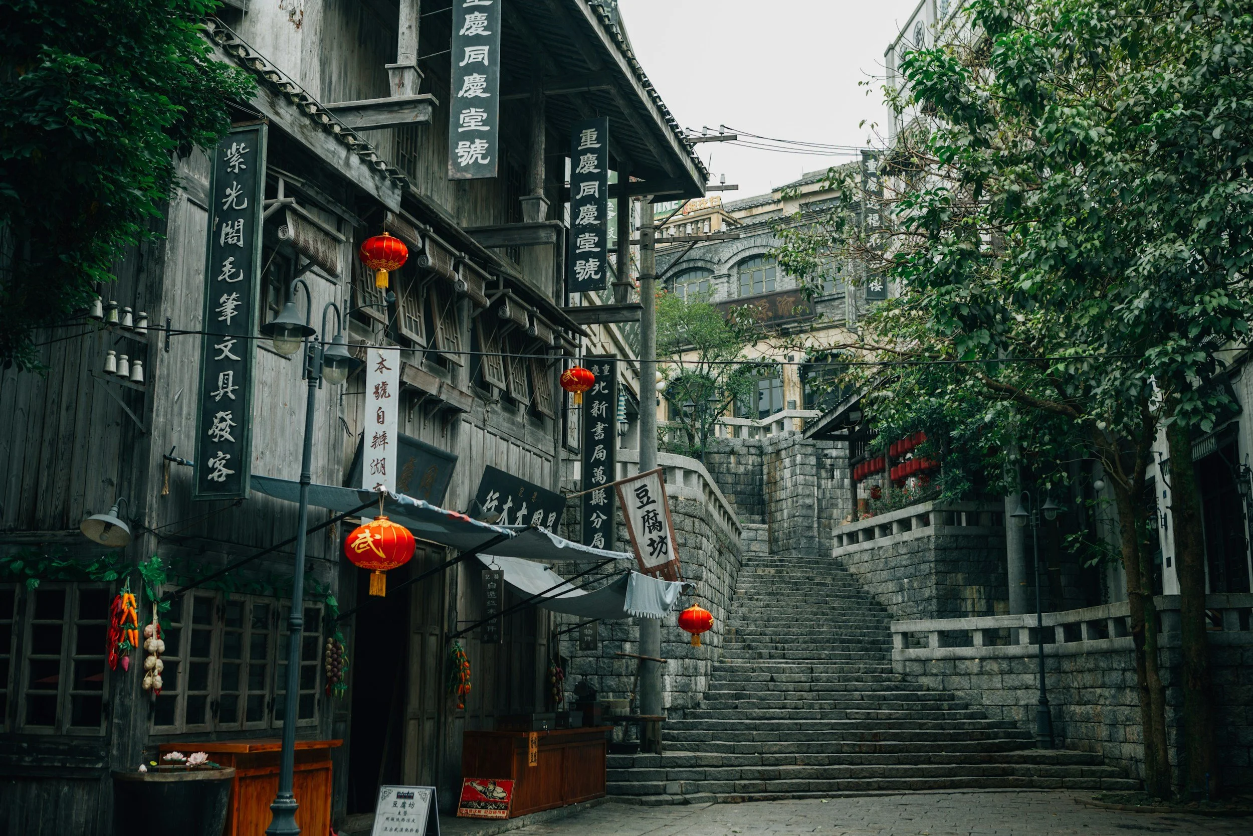 A traditional Asian street with wooden buildings, red lanterns, and stone steps leading uphill. Signs with Chinese characters are hanging on the buildings, and there are trees along the street.