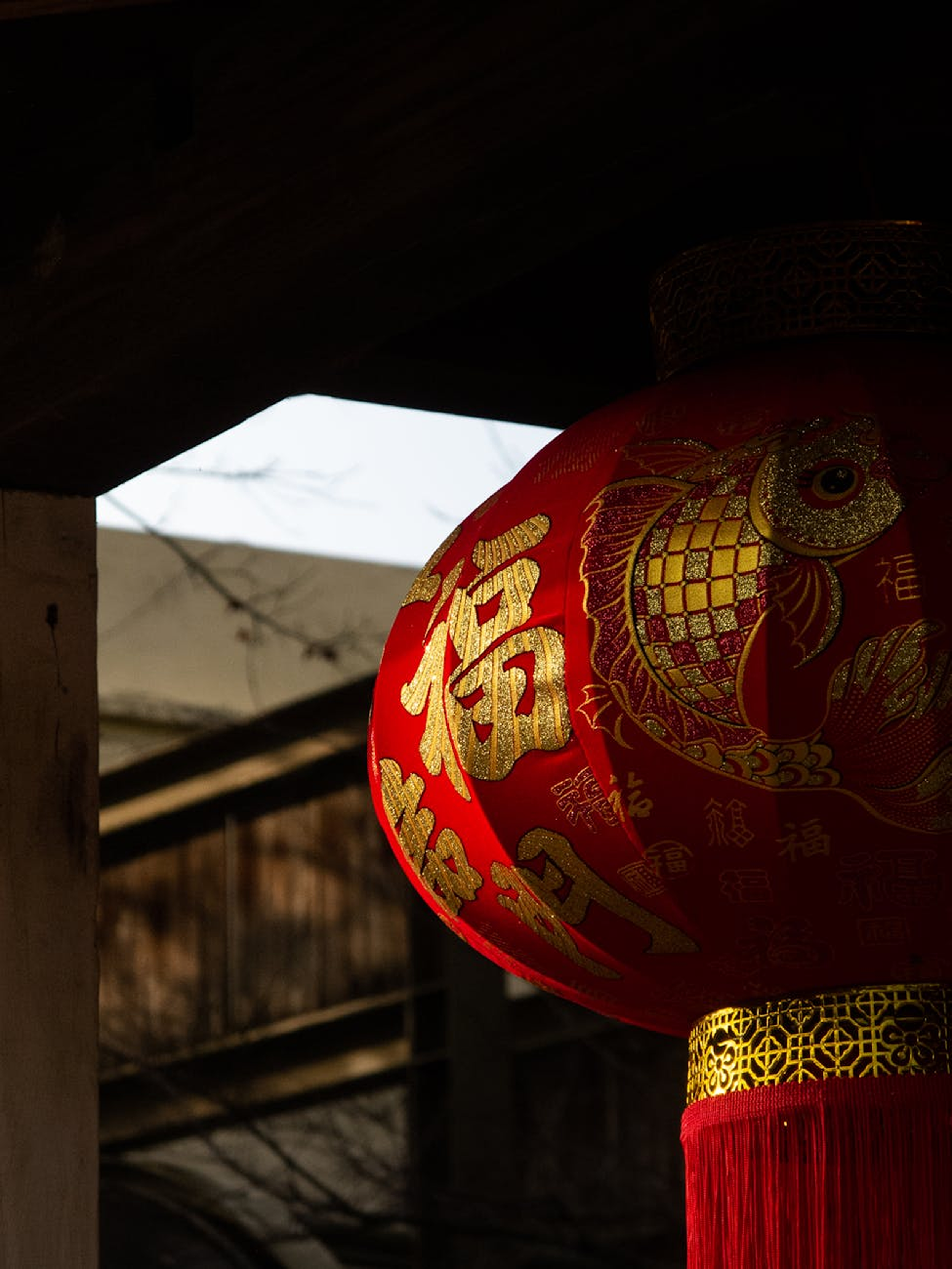 Close-up of a traditional Chinese red lantern with gold decorations and calligraphy, hanging in a shaded area.