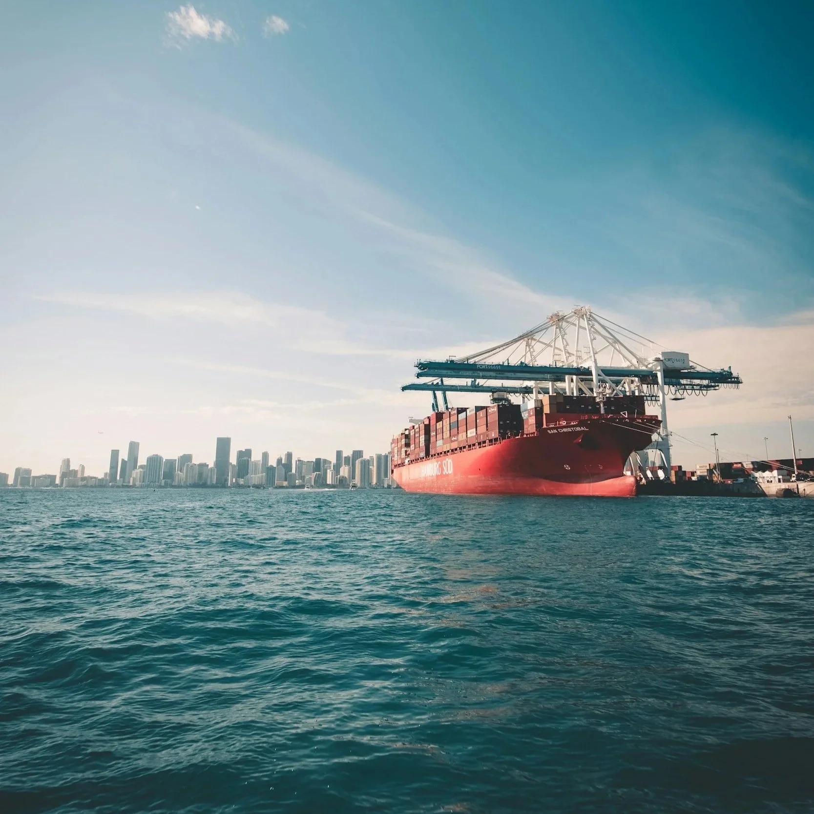 Large red cargo ship docked at a port with city skyline in the background, under a partly cloudy sky.
