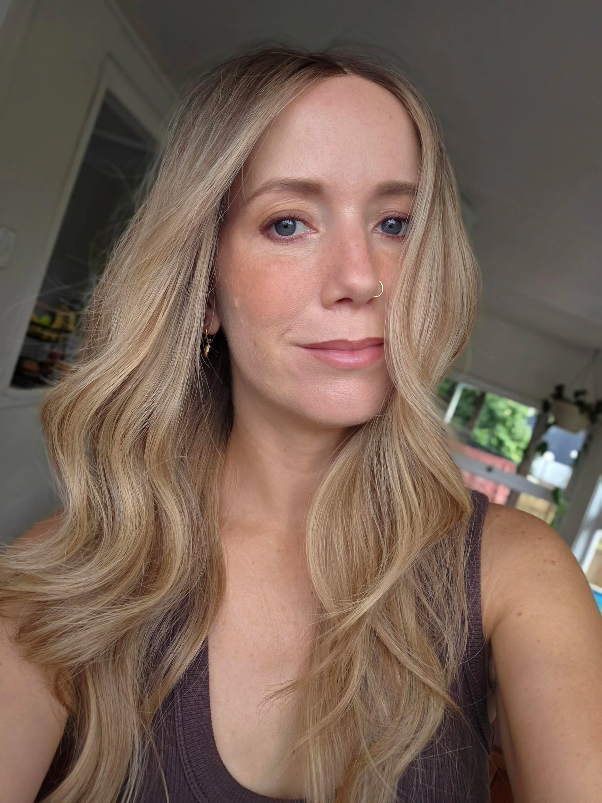 Close-up of a woman with long, wavy blonde hair, blue eyes, light makeup, a small nose ring, and wearing a sleeveless dark top, indoors with a window and some greenery visible in the background.