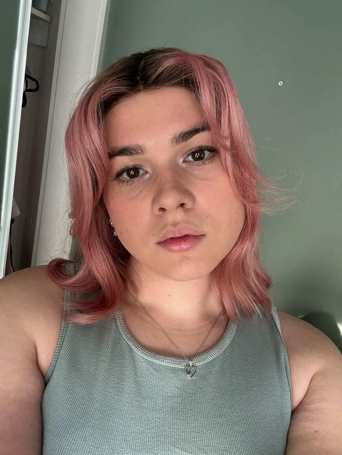 A young woman with pink hair and a silver heart-shaped necklace taking a selfie indoors, with a green wall and closet in the background.